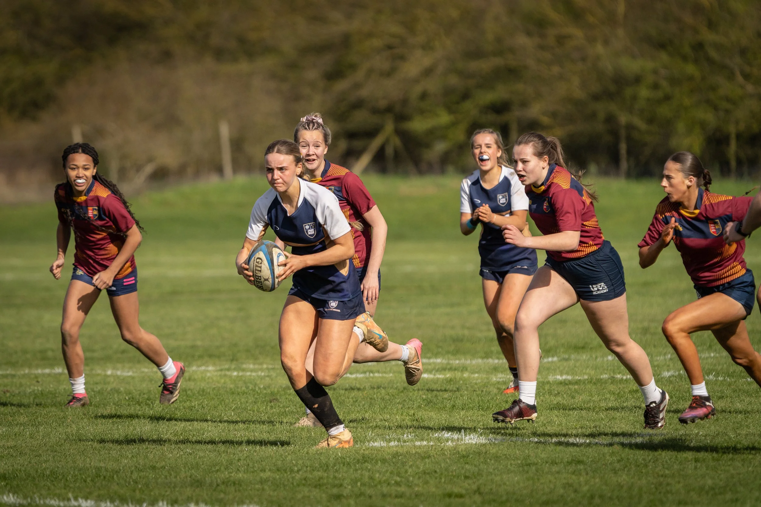 Young women playing rugby on a grassy field, wearing team jerseys, with some holding a rugby ball and others running.