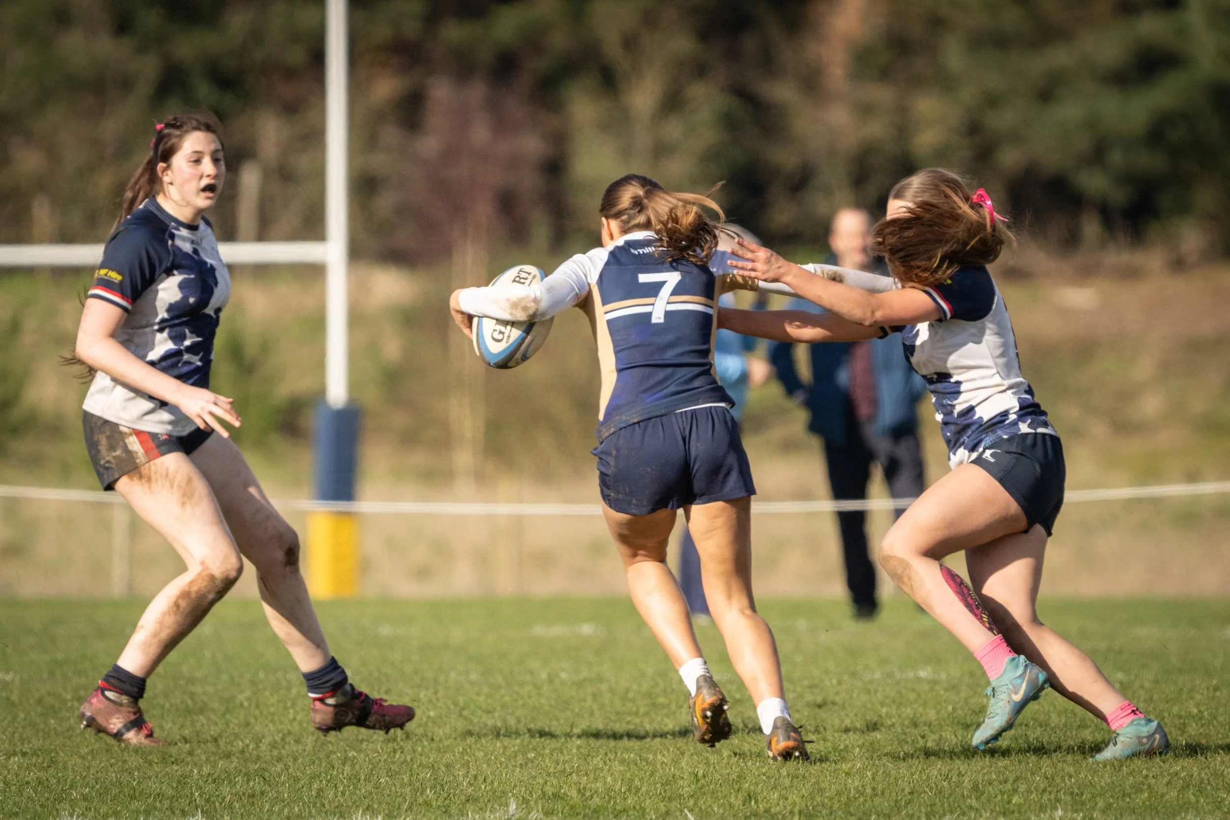 Girls playing rugby on a grassy field, with three girls in action fighting for possession of the rugby ball, one girl wearing black shorts and two in blue and white jerseys.