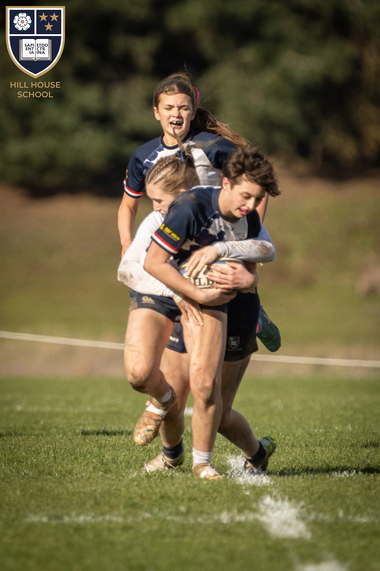 A rugby game with three young players, one carrying the ball, on a grassy field. Two players are marking the ball carrier, with one behind her and the other nearby. All players are wearing rugby uniforms and cleats.