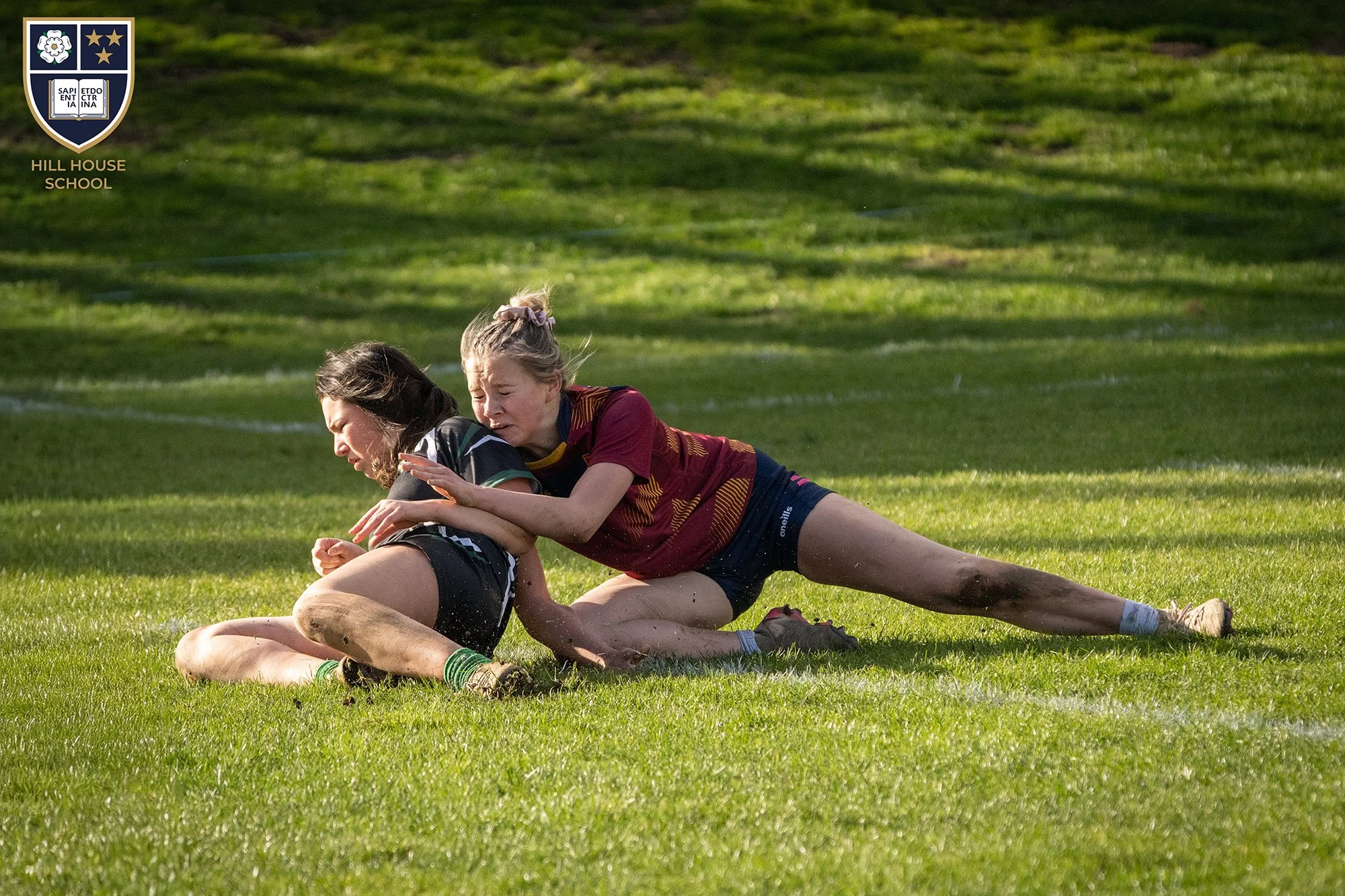 Two young women wrestling on a grassy field, one wearing a maroon and yellow sports jersey and the other in a black and green athletic shirt. They are engaged in a competitive match, with one woman pinning the other down.