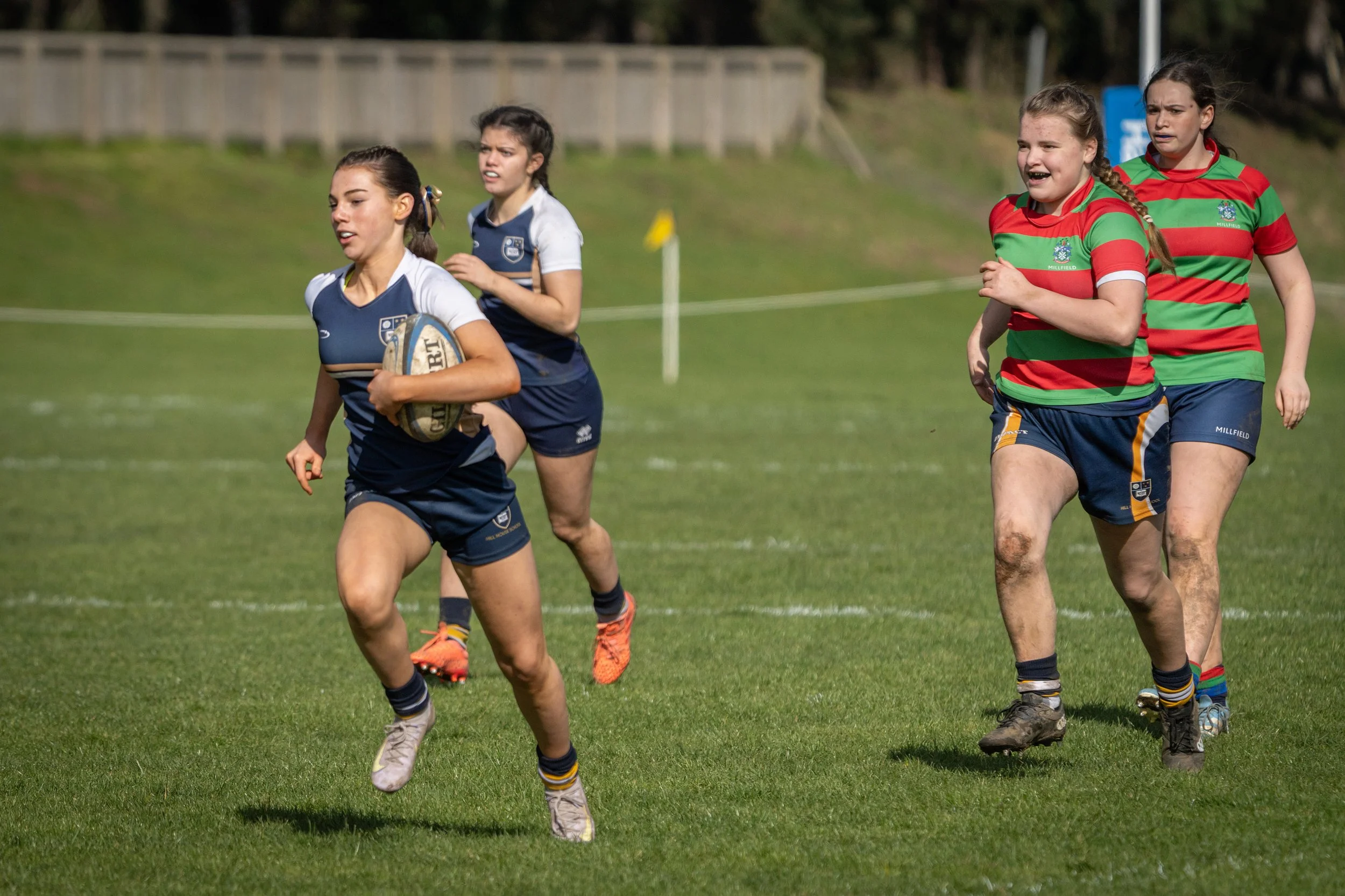 Four young female rugby players on a grassy field, wearing sports uniforms, running during a game.