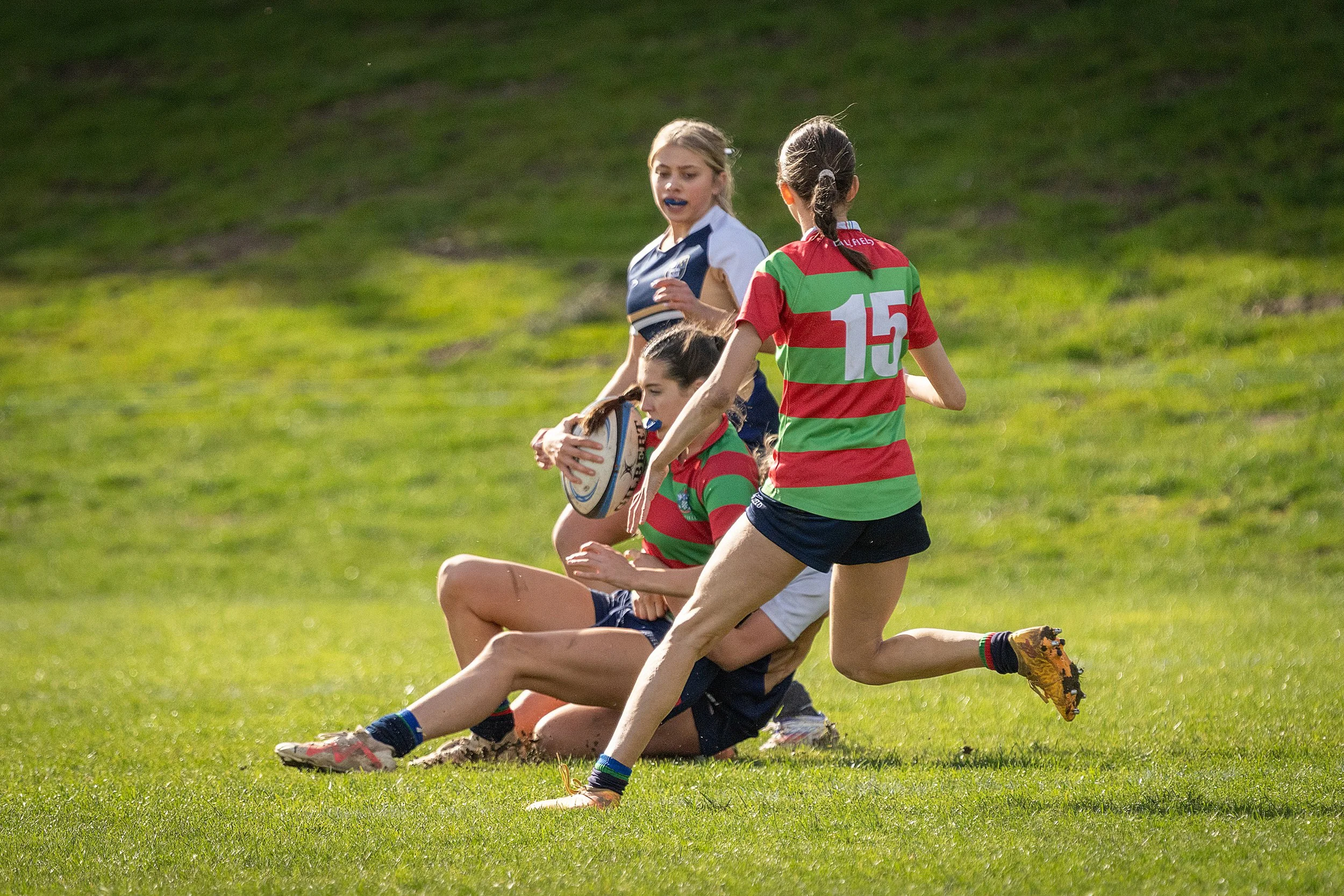 Young girls playing rugby outdoors on a grassy field, with some players wearing uniforms in red and green stripes and others in blue and white.