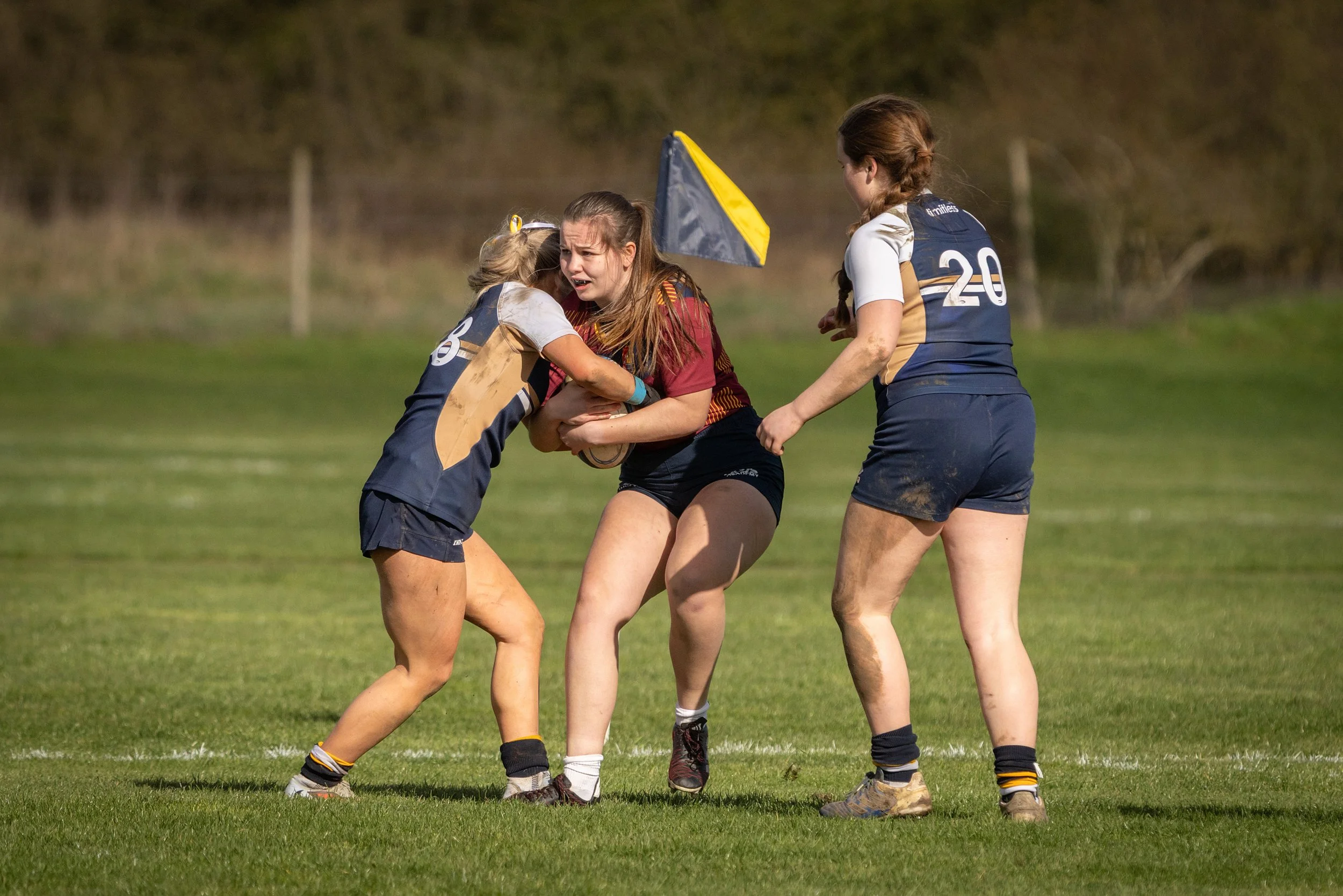 Girls playing rugby on a grassy field, with one girl holding a rugby ball and others in a tackle, wearing sports uniforms.