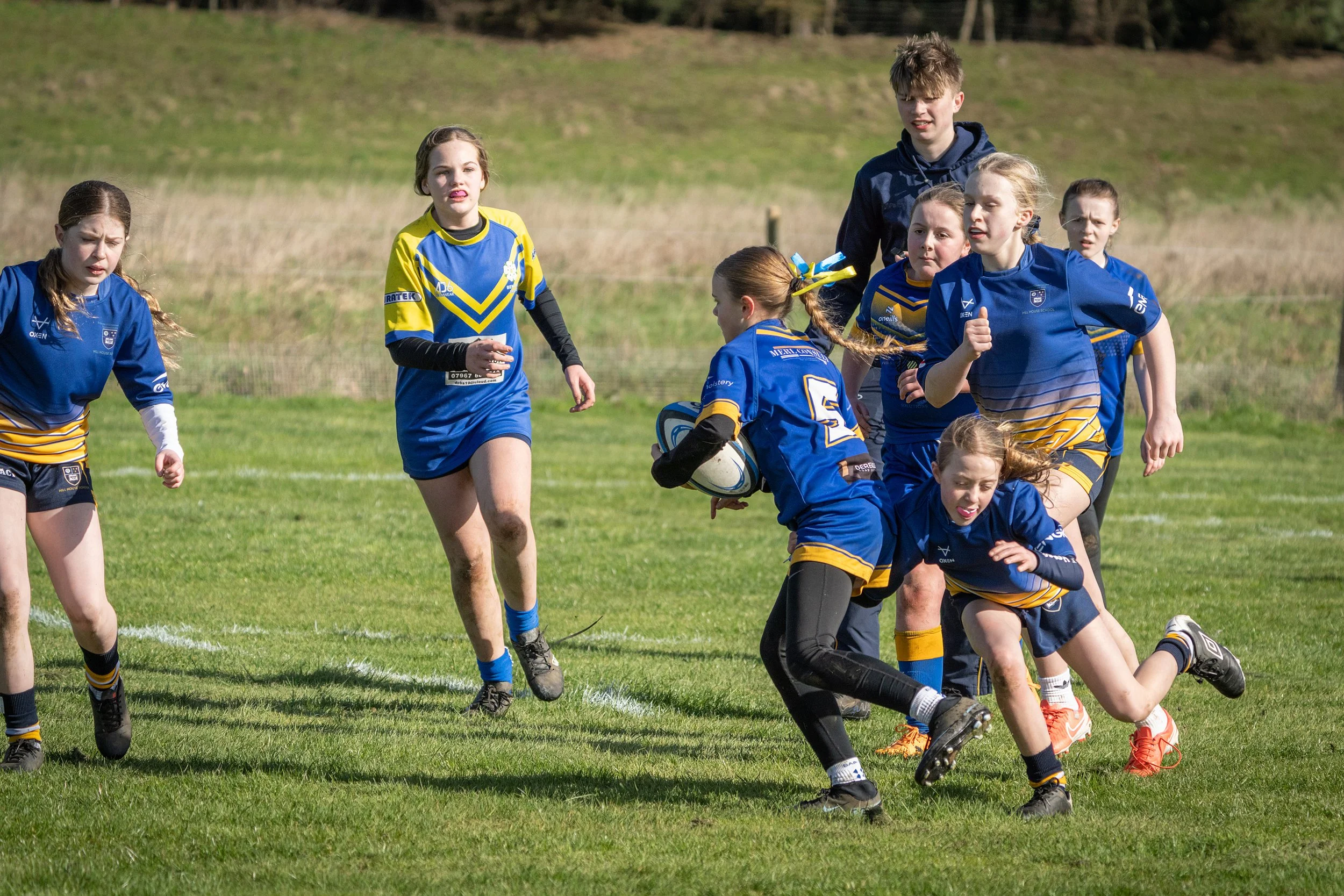 Children playing rugby on a grassy field, wearing blue and yellow uniforms, with a few players on the ground and one holding a rugby ball.