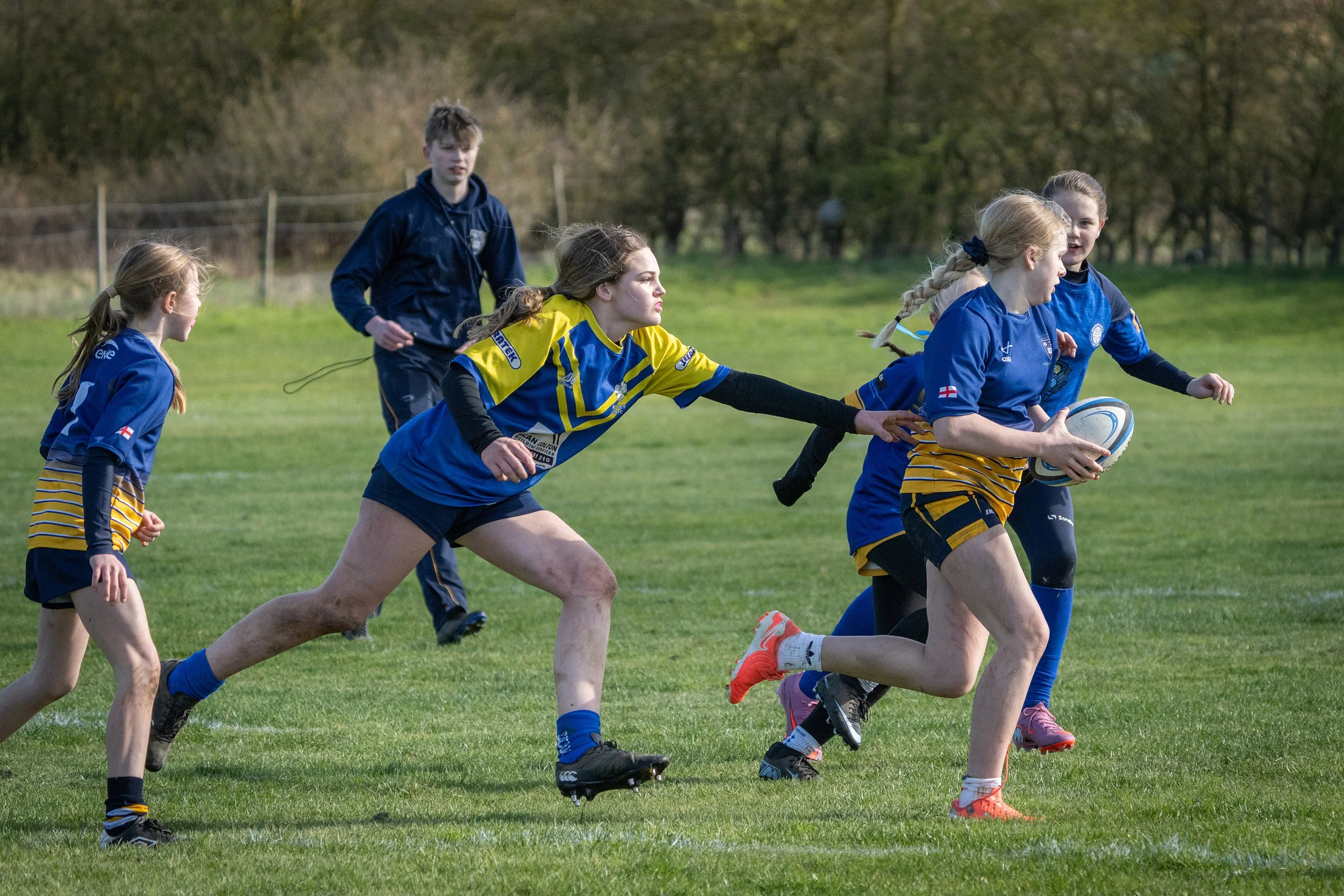 Girls playing rugby on a grassy field with trees in the background.