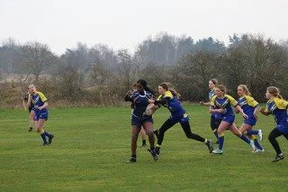 A group of young women playing rugby on a grassy field, with trees in the background.
