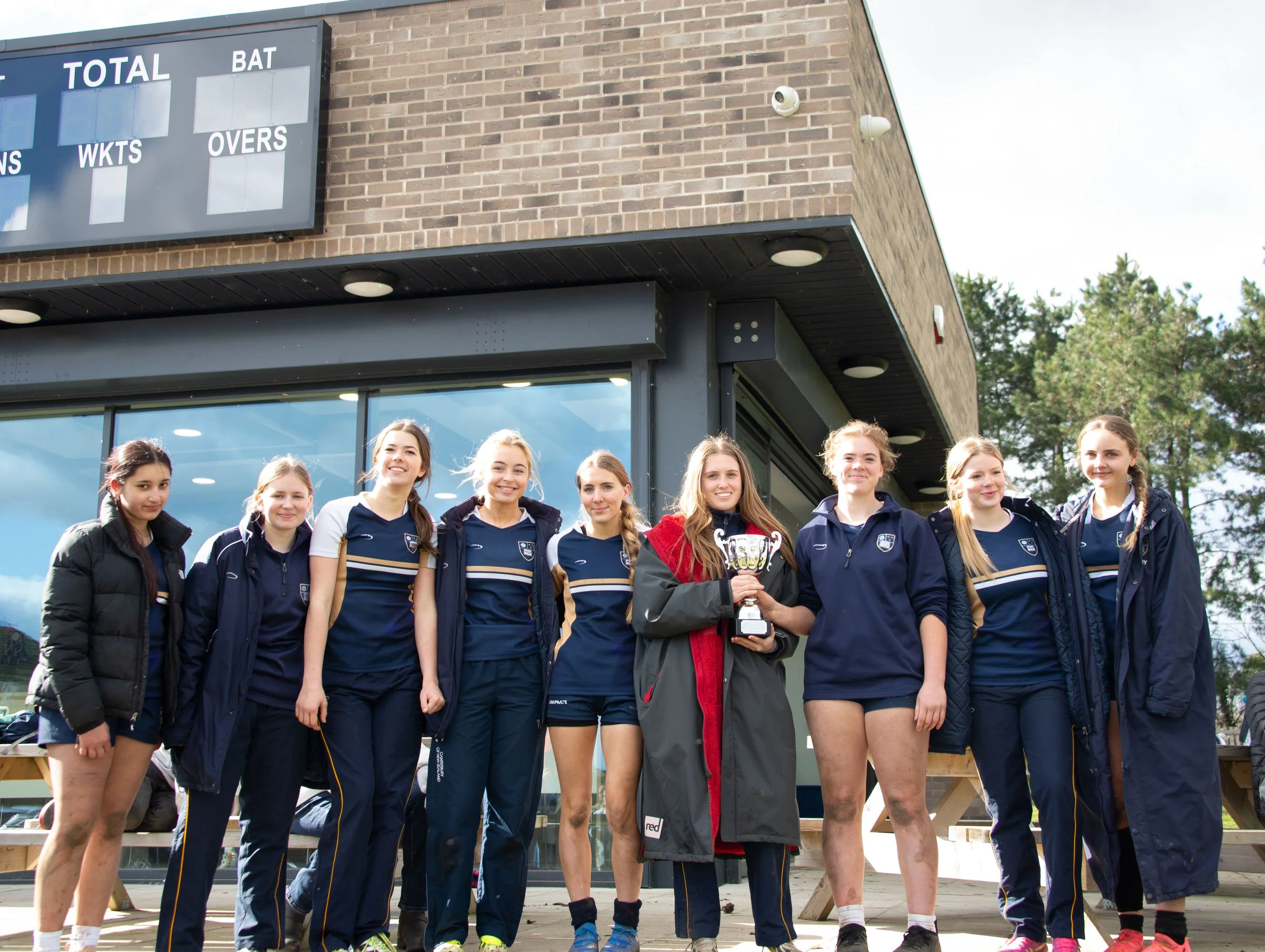 A group of young female athletes standing together outdoors in front of a scoreboard, with one holding a trophy, all wearing navy blue sports uniforms, smiling.
