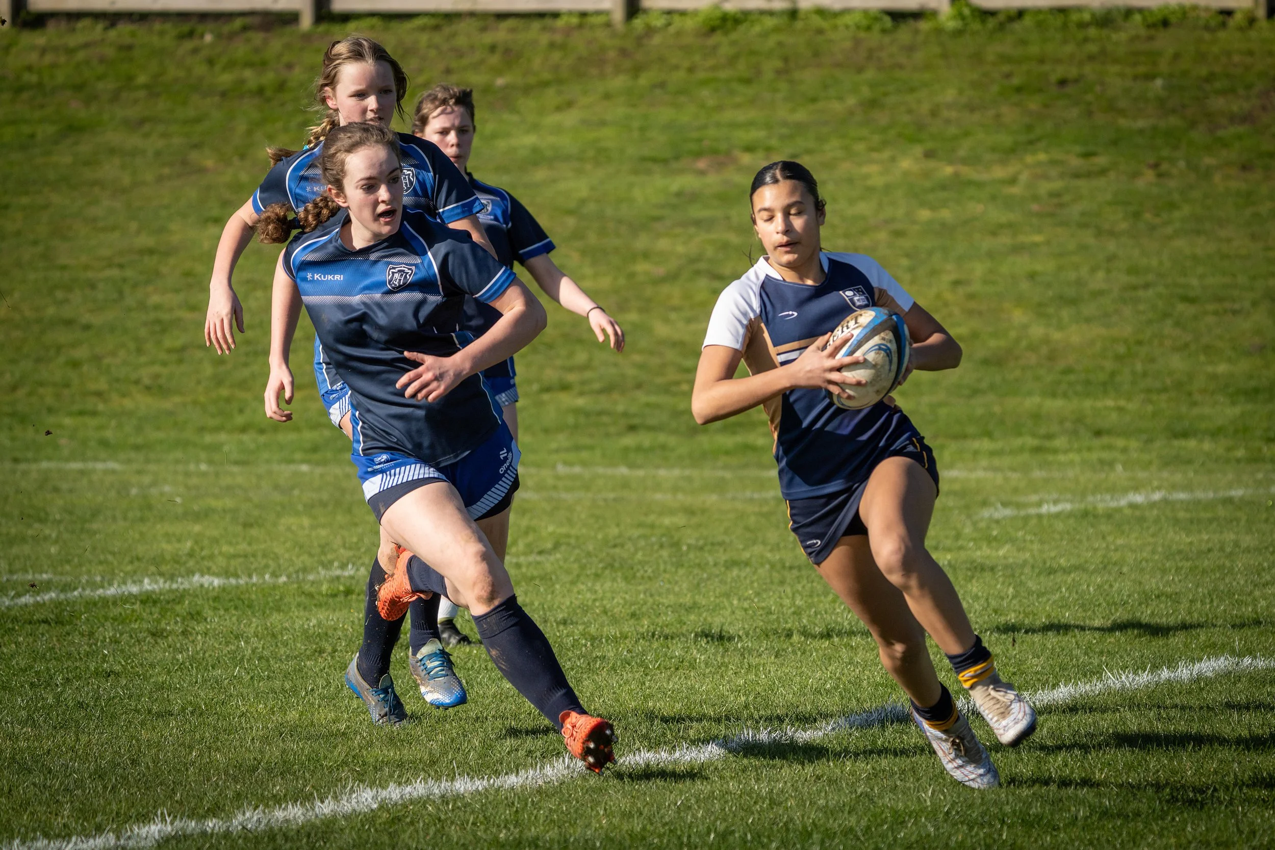 A group of female rugby players running on a grassy field, with one player holding a rugby ball, wearing uniforms in navy blue and white.