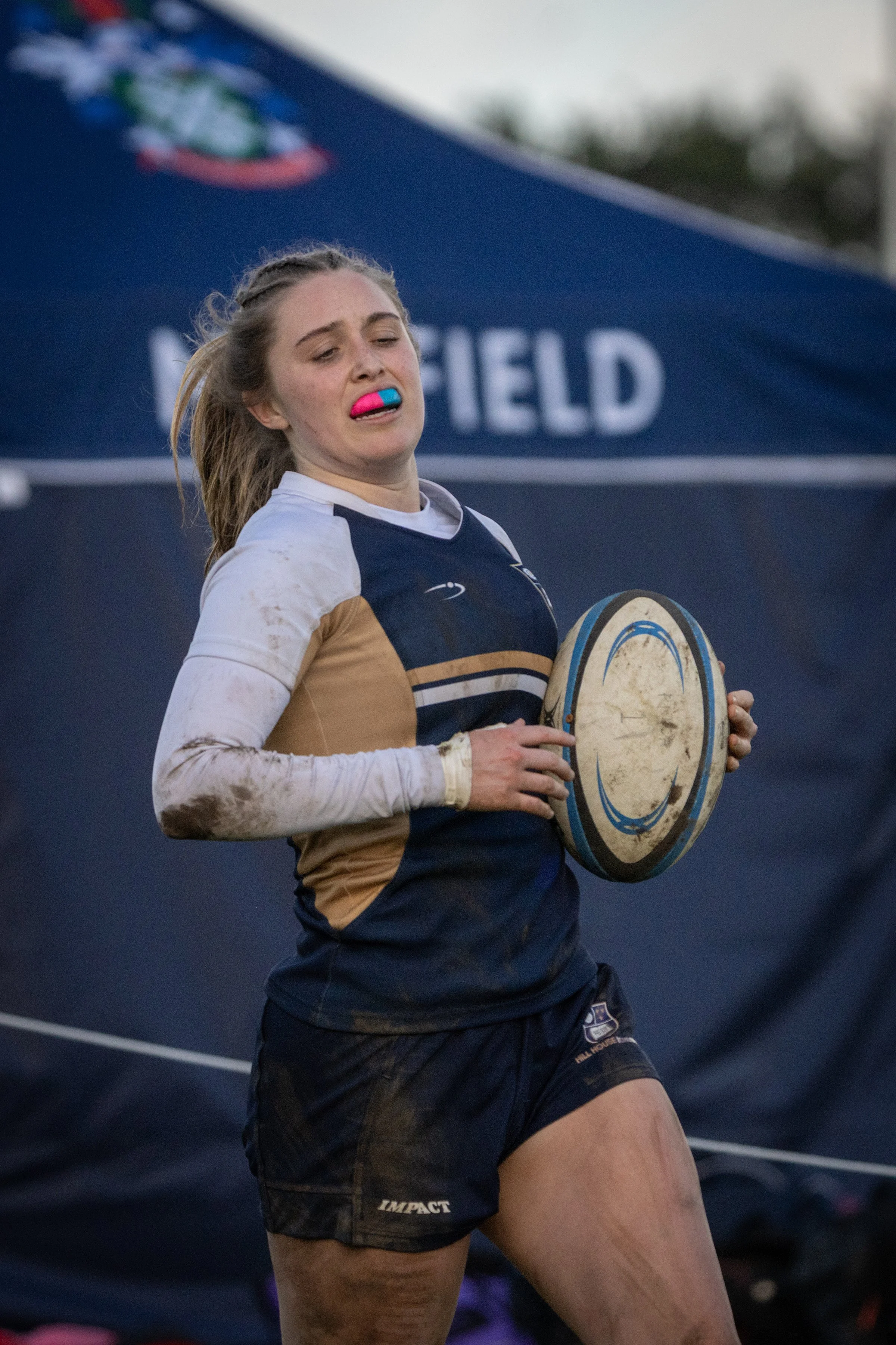 A female rugby player holding a rugby ball, wearing a navy and tan jersey, with mud on her sleeves and face, standing in front of a blue tent at a rugby field.