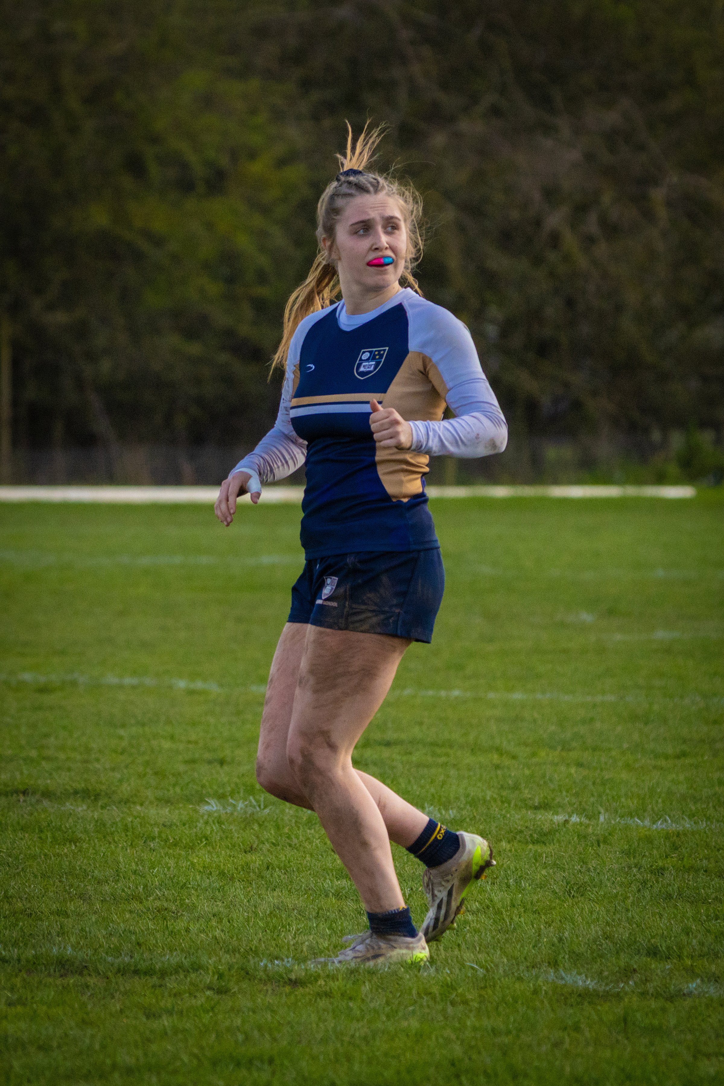 A female rugby player running on a grassy field during a game, wearing a blue and tan jersey with a crest on the chest, black shorts, and rugby cleats, with dirt and mud on her legs.