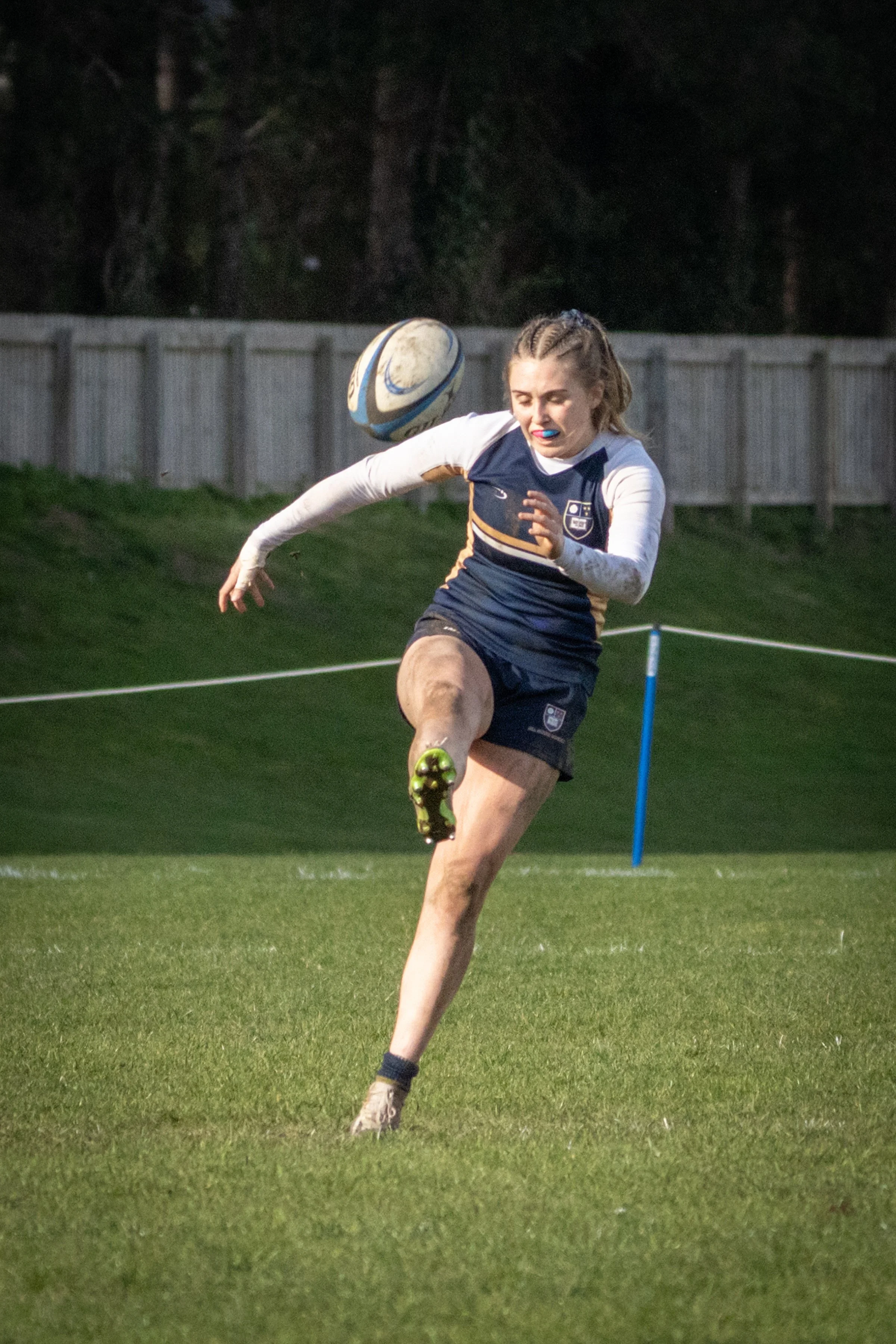A female rugby player running on a grassy field during a game, wearing a blue and white uniform, with a rugby ball in mid-air near her head, and a fence in the background.