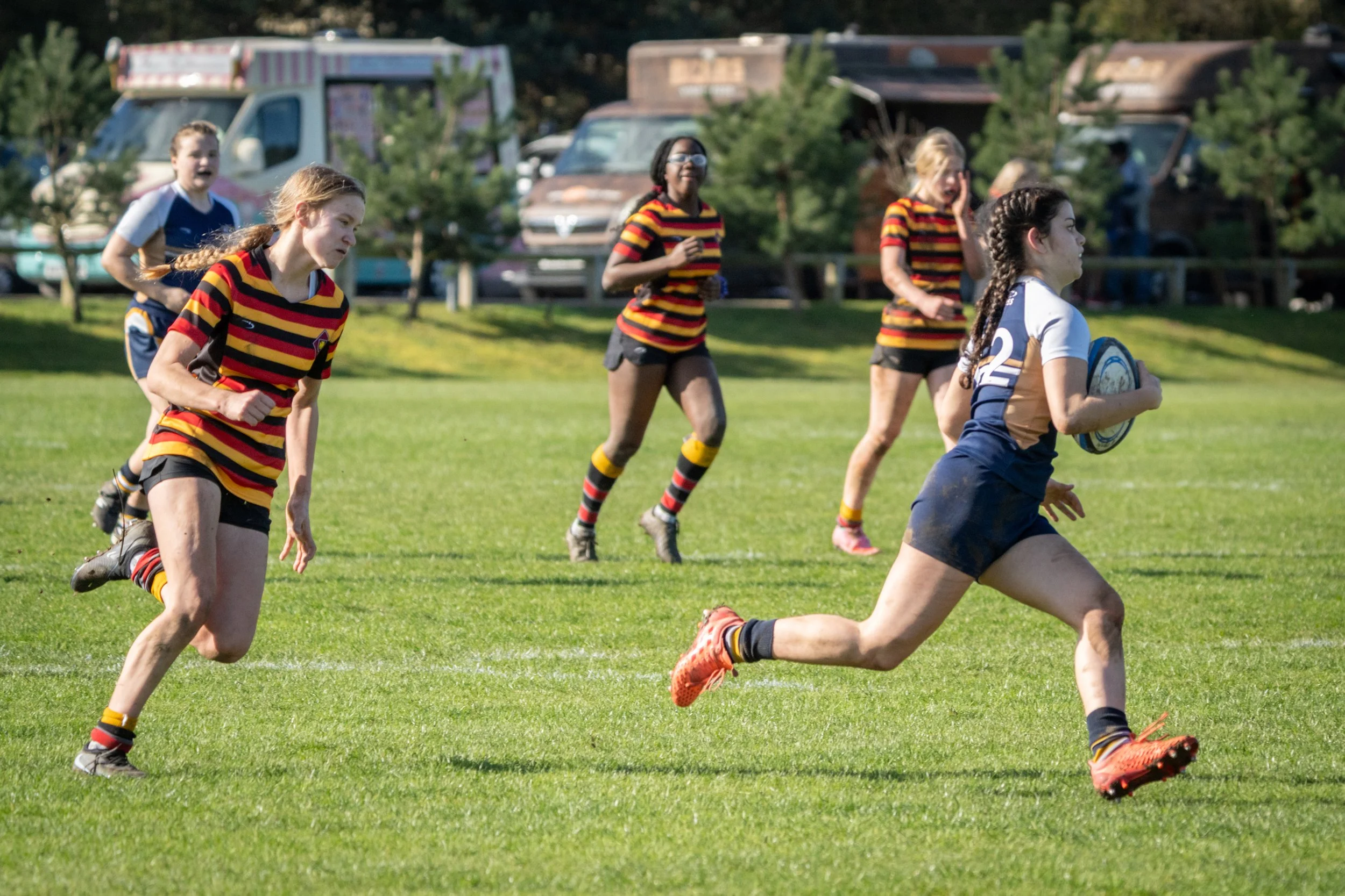 Women and girls playing rugby on a grassy field with trees and parked vehicles in the background.