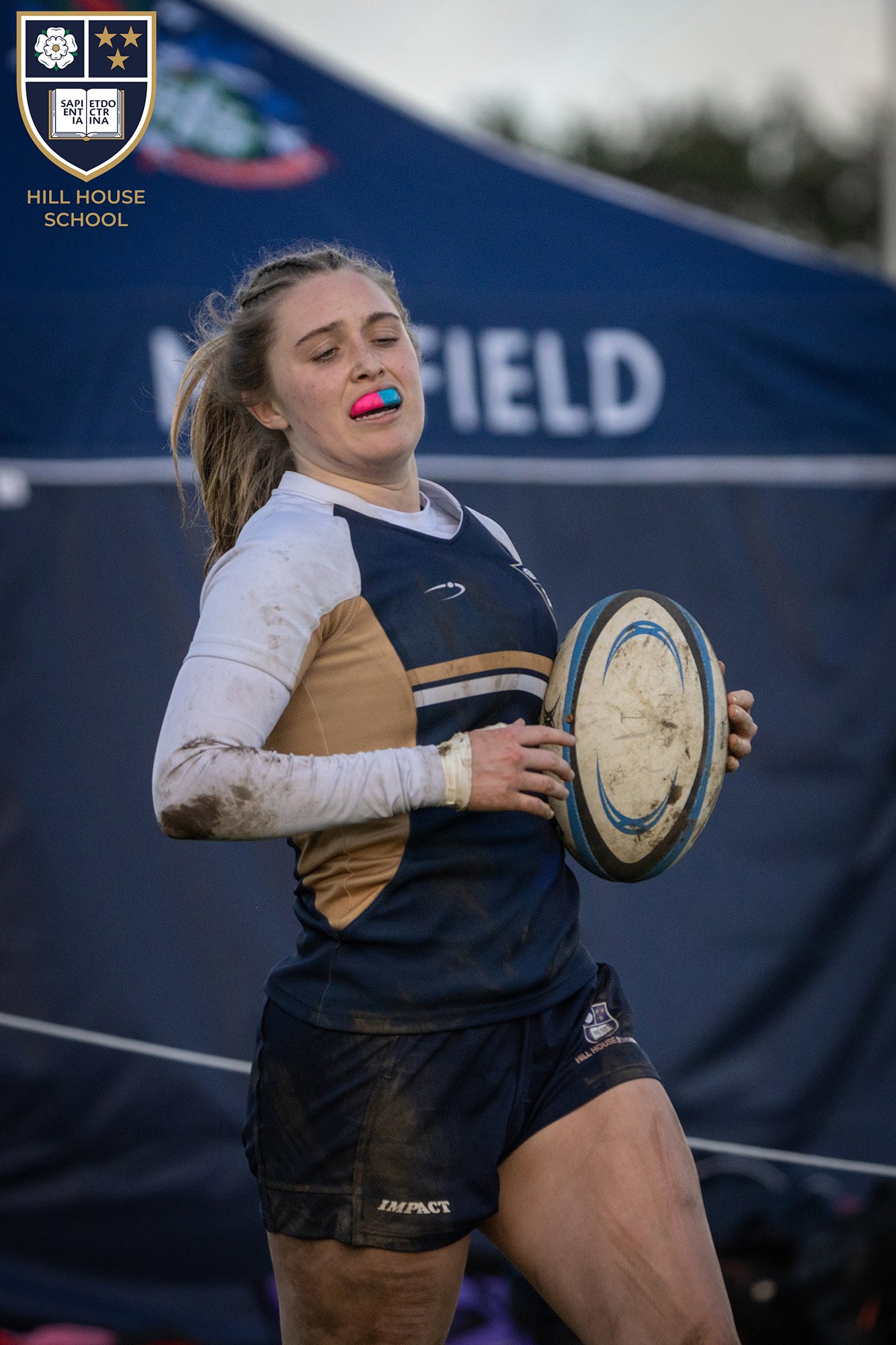 A female rugby player with a pink and blue mouthguard, holding a rugby ball, wearing a navy and beige sports uniform, appears exhausted or strained, with a muddy arm visible in the background, on a rugby field.