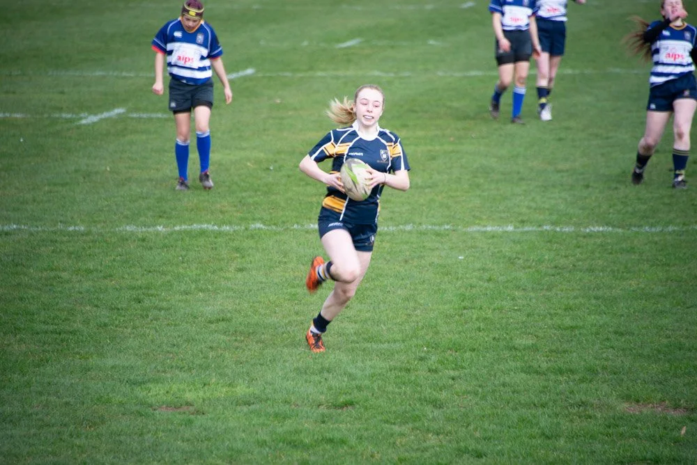 A young girl playing rugby, running with a rugby ball on a green field, with other children in the background.