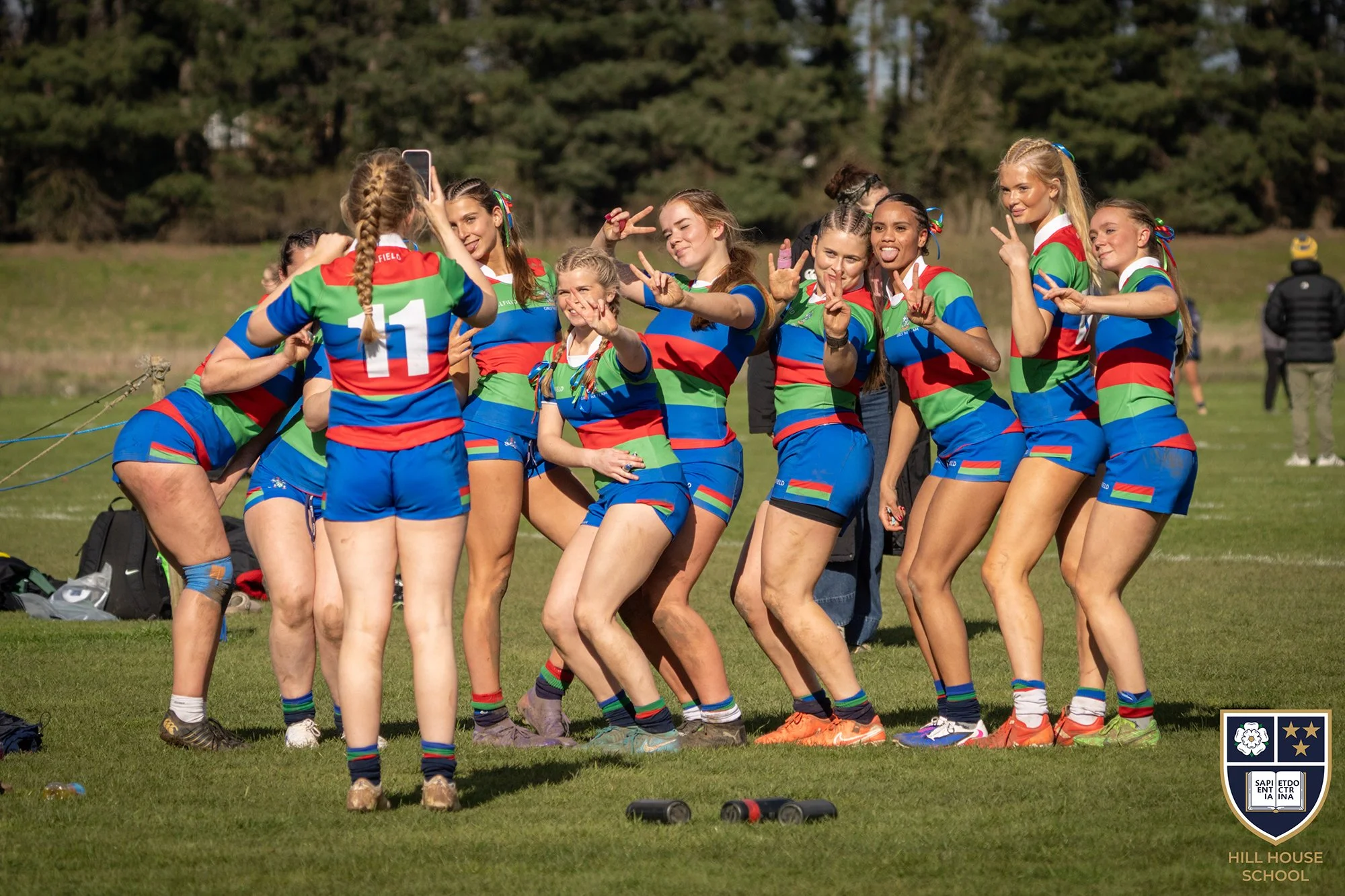 A group of female rugby players wearing colorful uniforms taking a selfie on a grassy field, with a few onlookers and trees in the background.
