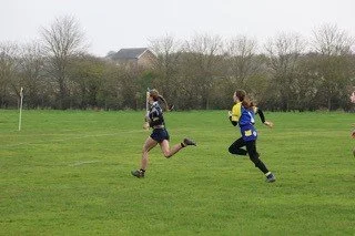 Two girls running on a grassy field during daytime, with trees and houses in the background.