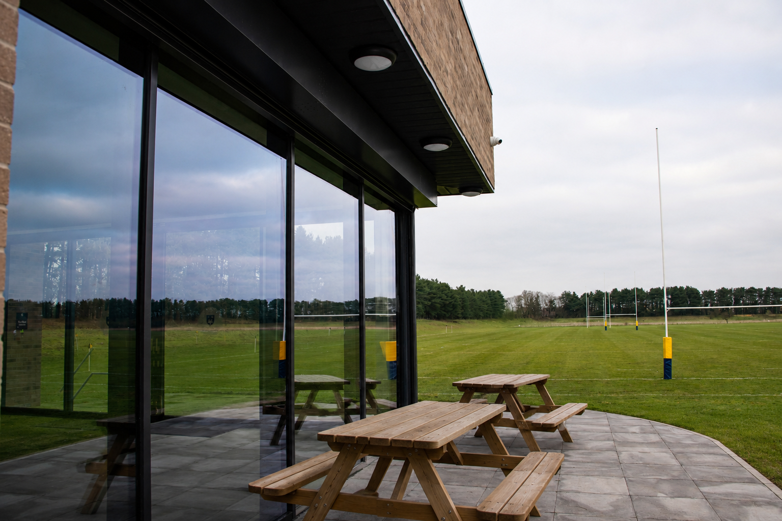 Outdoor patio area with wooden picnic tables next to a building with large glass windows, overlooking a rugby field with goalposts under a cloudy sky.