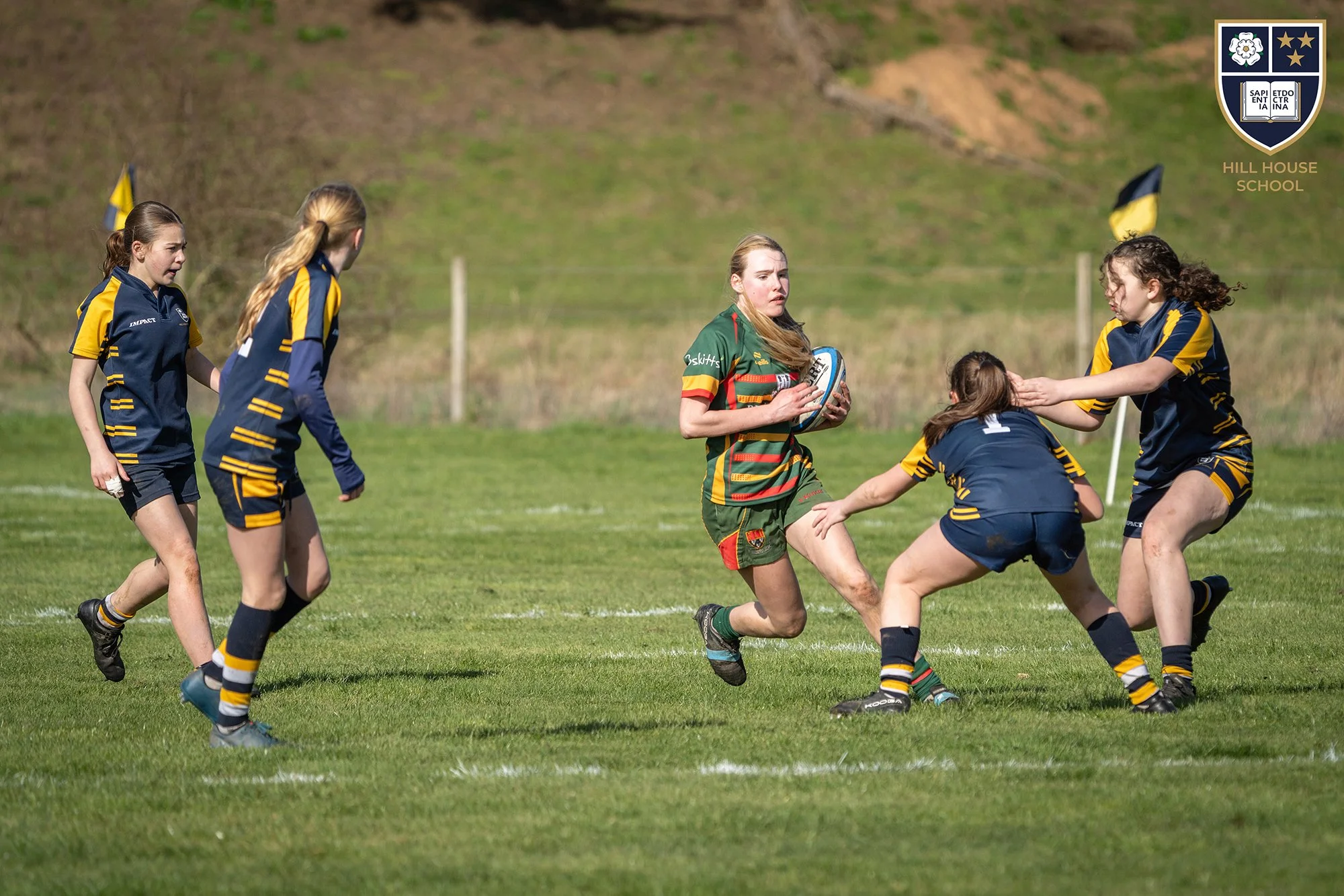 Girls playing rugby on a grassy field, with a hill in the background and a school crest logo in the top right corner.