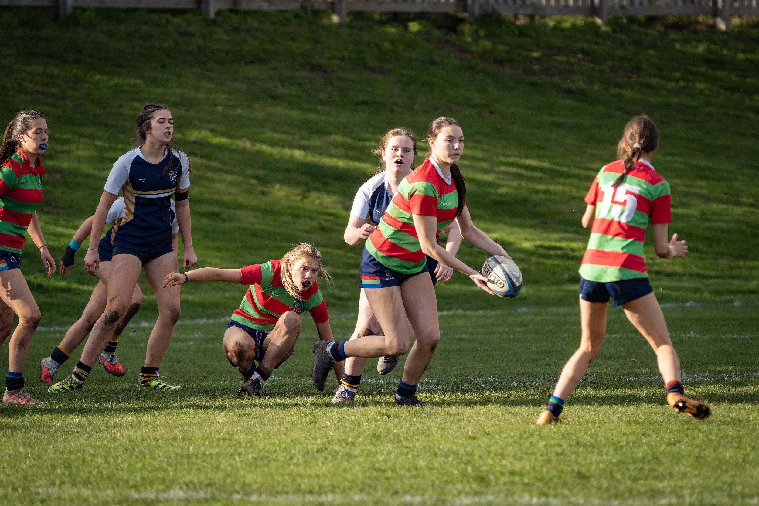 A group of young women playing rugby on a field, wearing colorful striped jerseys and shorts.