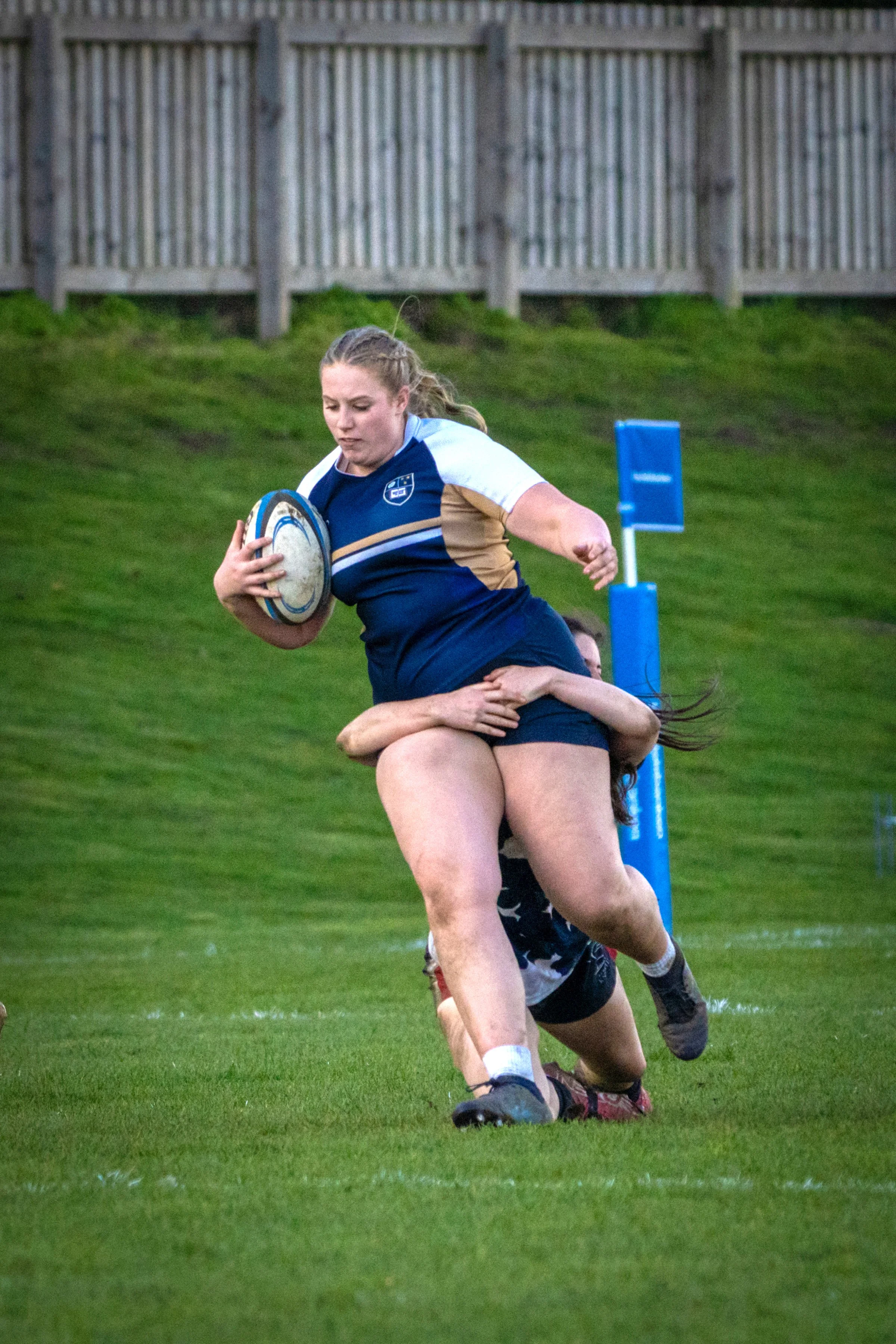 A female rugby player in a blue and gold uniform carrying a rugby ball while running on a grassy field, with opponents attempting to tackle her.