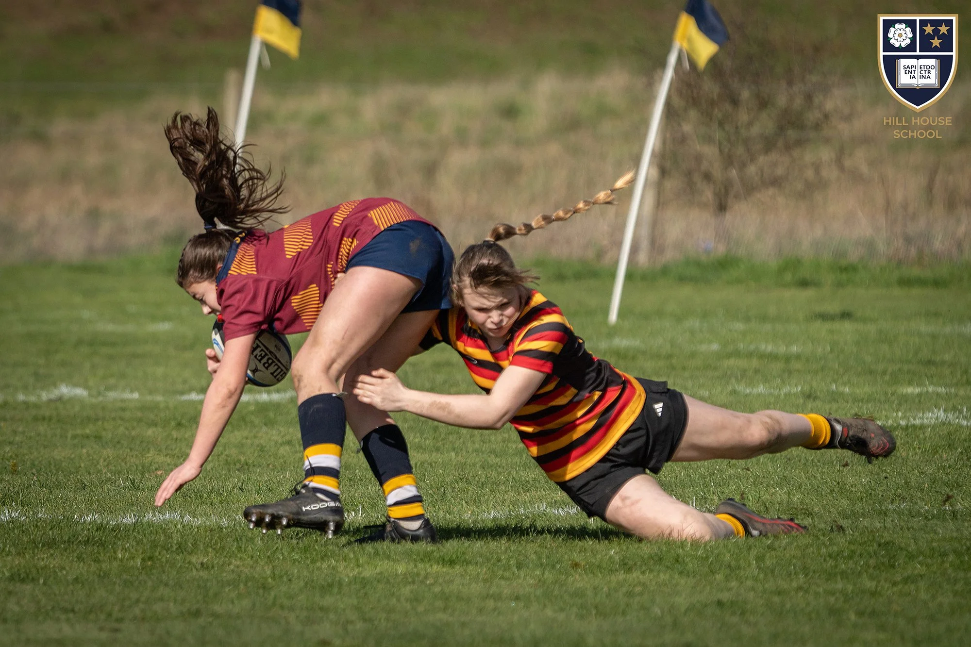 Two rugby players, one in a maroon and yellow jersey and the other in a striped black, red, and yellow jersey, are engaged in a tackle on the field, with flags in the background and a school emblem in the upper right corner.