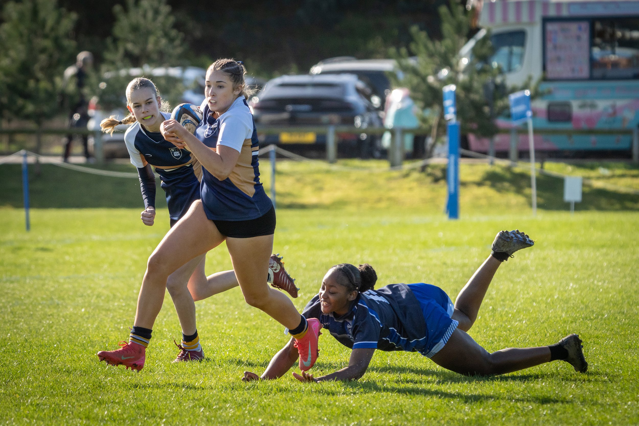 Two female rugby players wearing blue and gold uniforms are competing on a grassy field. One is running with the ball while the other is attempting to tackle her, and a third player is on the ground. In the background, there are cars and a colorful b