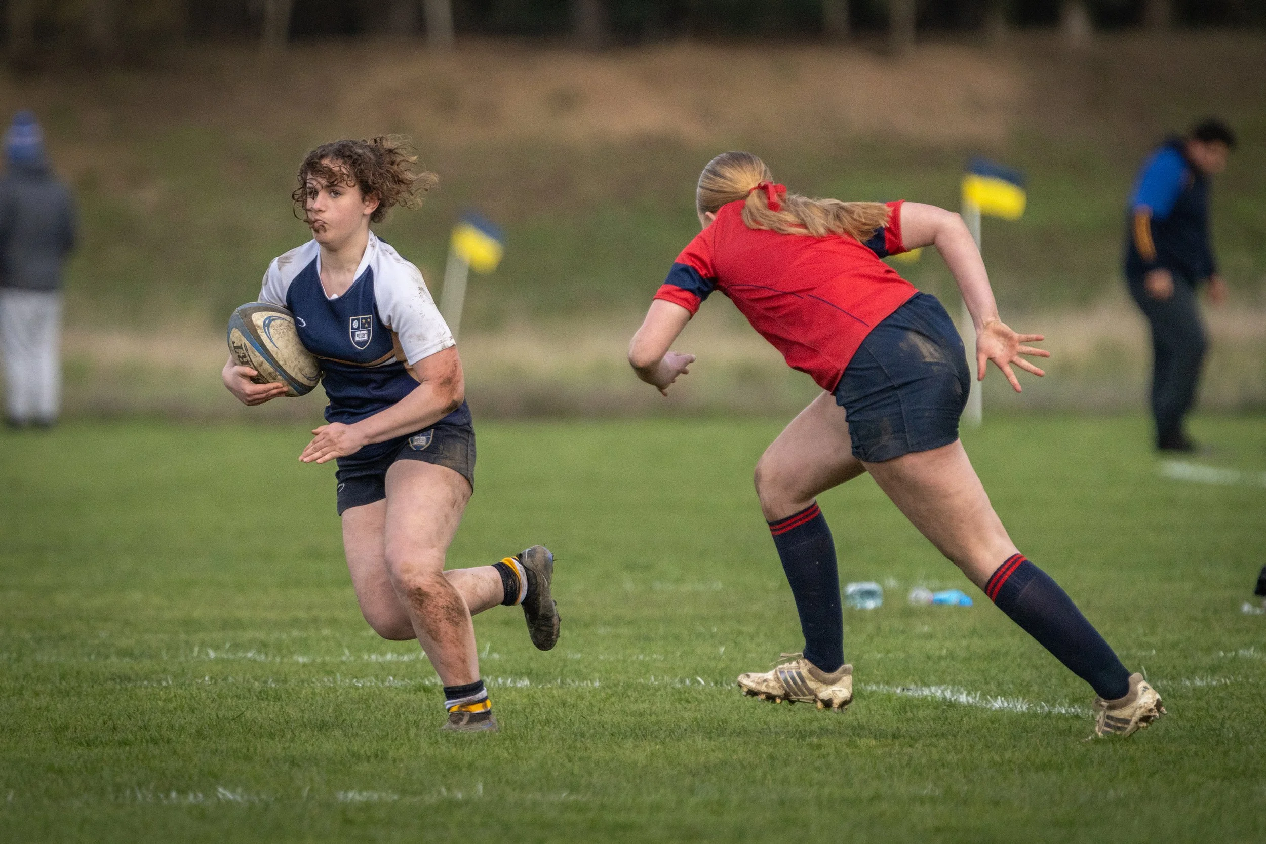 Two young female rugby players in action on a grassy field; one in a blue and white jersey holding a rugby ball, the other in a red jersey attempting to tackle.
