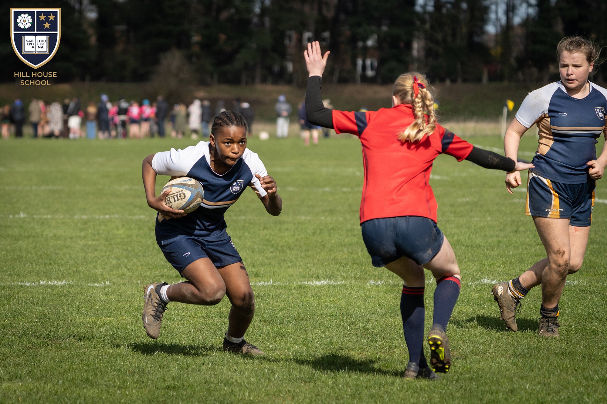 Girls playing rugby on a grassy field, with one girl running with the ball and another girl in a red jersey attempting to block her, while a third girl looks on. In the background, people are watching the game.