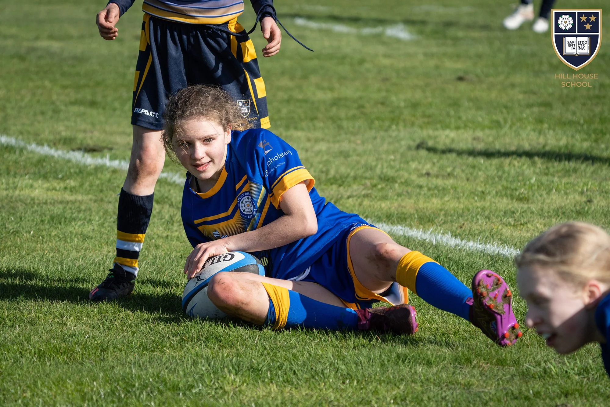 A young girl in a blue and yellow rugby uniform lying on the grass holding a rugby ball, with another person standing nearby on a sports field with white lines and a school crest in the corner.