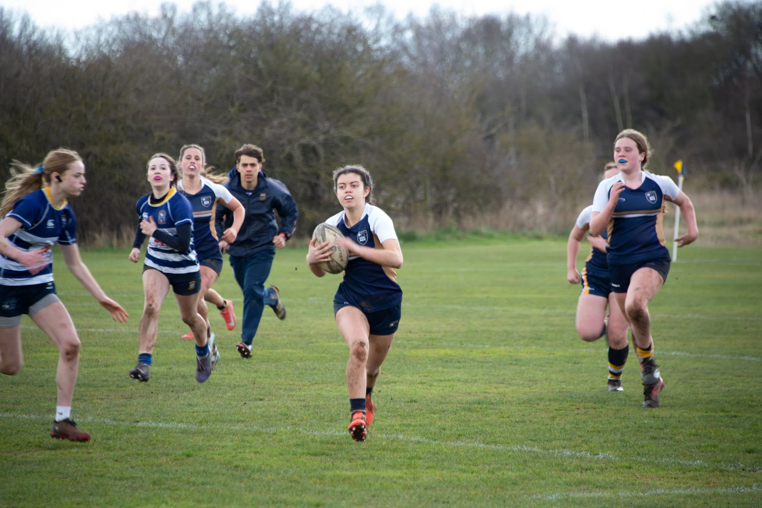A group of young women and one young man playing rugby on a grassy field during daytime. The players are running towards the camera, with the girl in the center holding a rugby ball. They are dressed in sports uniforms, mostly navy blue and white, an