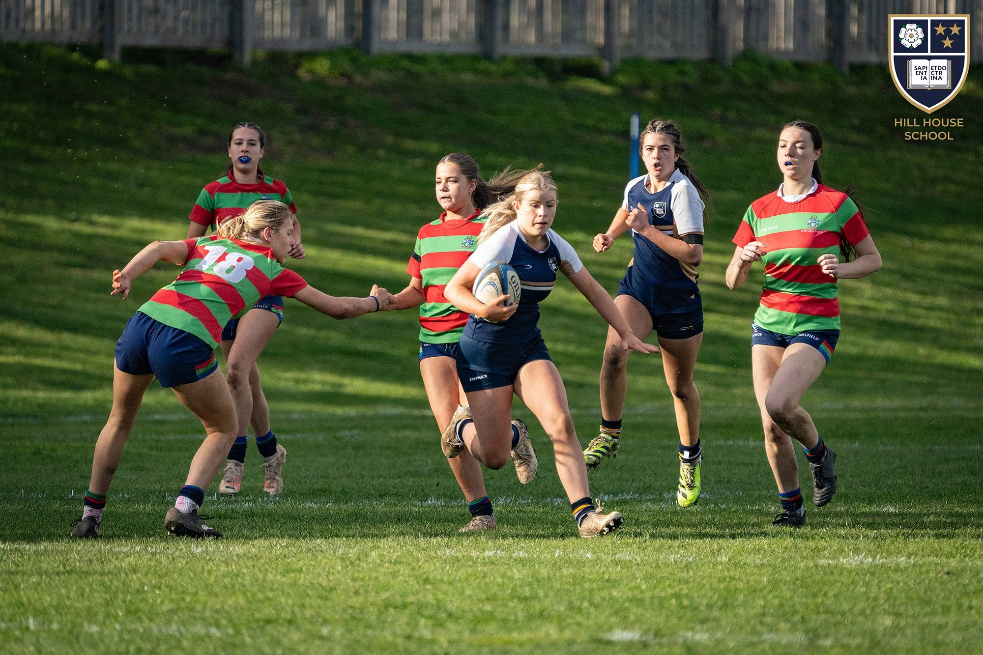 Girls playing rugby on a grassy field, one girl holding a rugby ball and others running around.