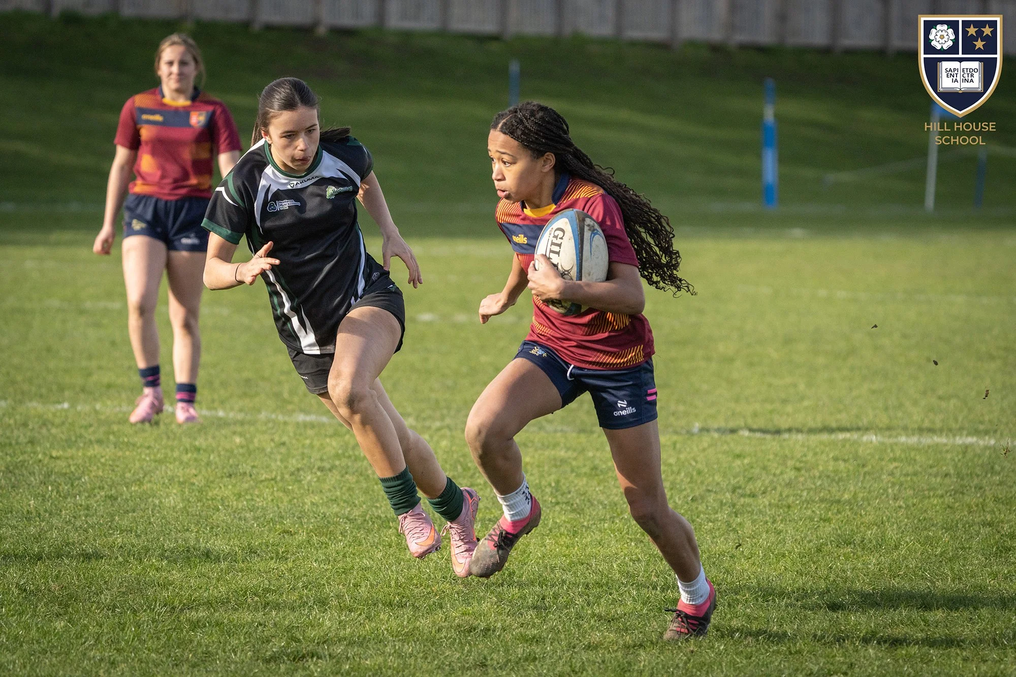 A girls' rugby match on a grassy field with two players competing for the ball. One player in a maroon and yellow jersey holds the ball, while the other in a black and green jersey attempts to block her. A third player in the background wears a maroo
