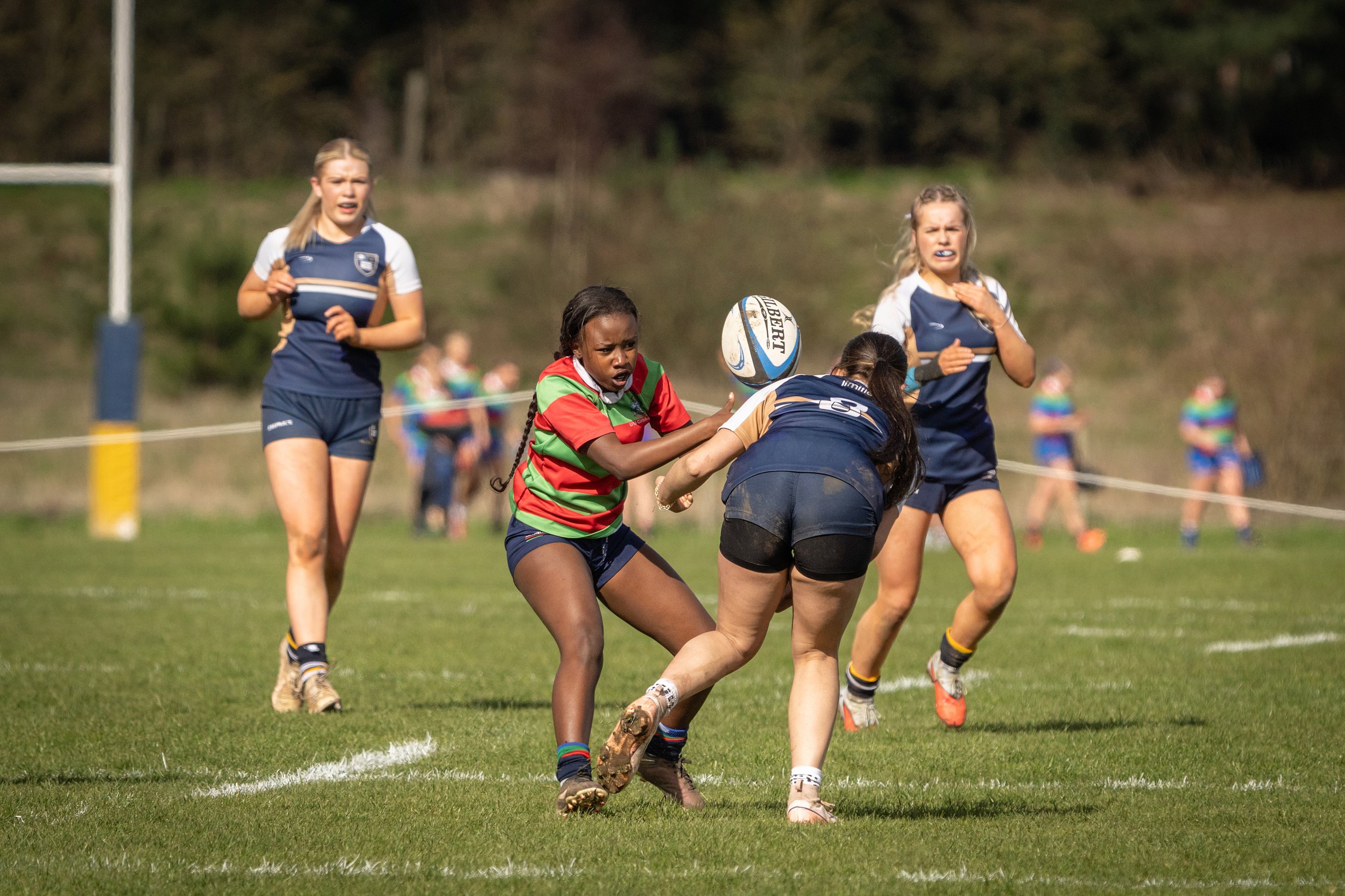 Women and girls playing rugby on a grassy field during daytime, with some players in blue uniforms and one in a striped red and green uniform.