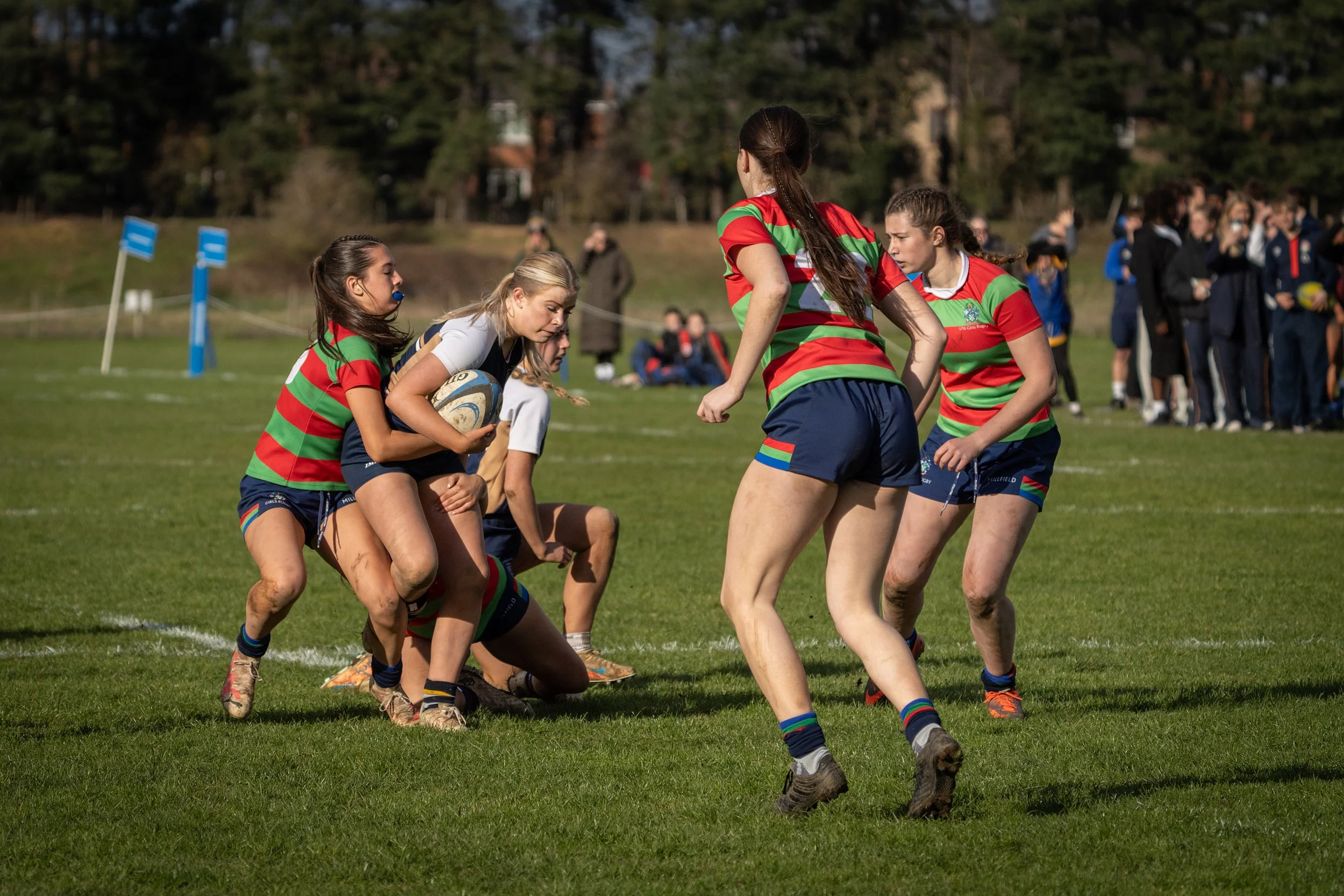 Women playing rugby on a grassy field, with one player holding a rugby ball, others locking in a scrum, and spectators in the background.