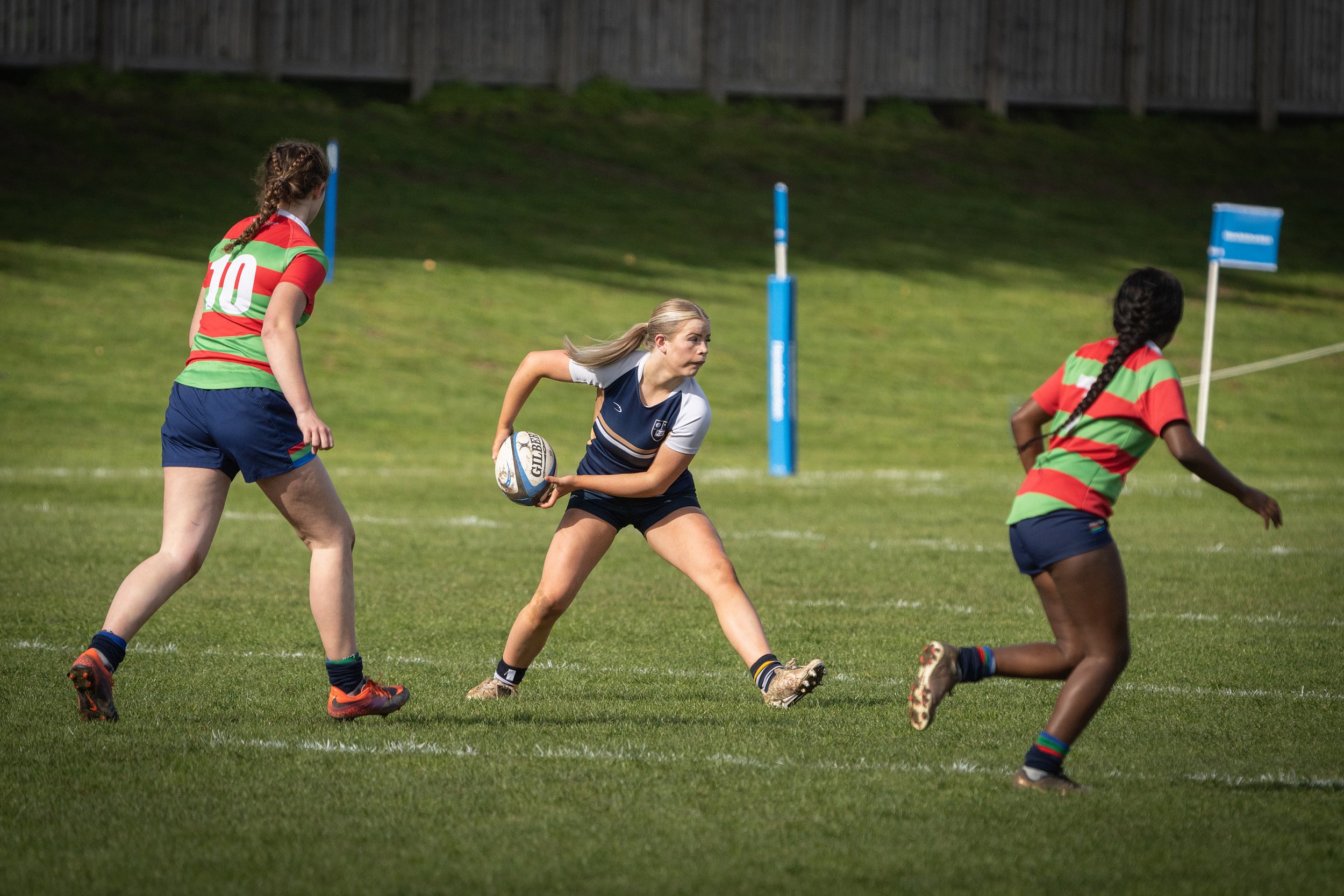 Young women playing rugby on a grassy field during daytime, with one holding a rugby ball and others moving around.