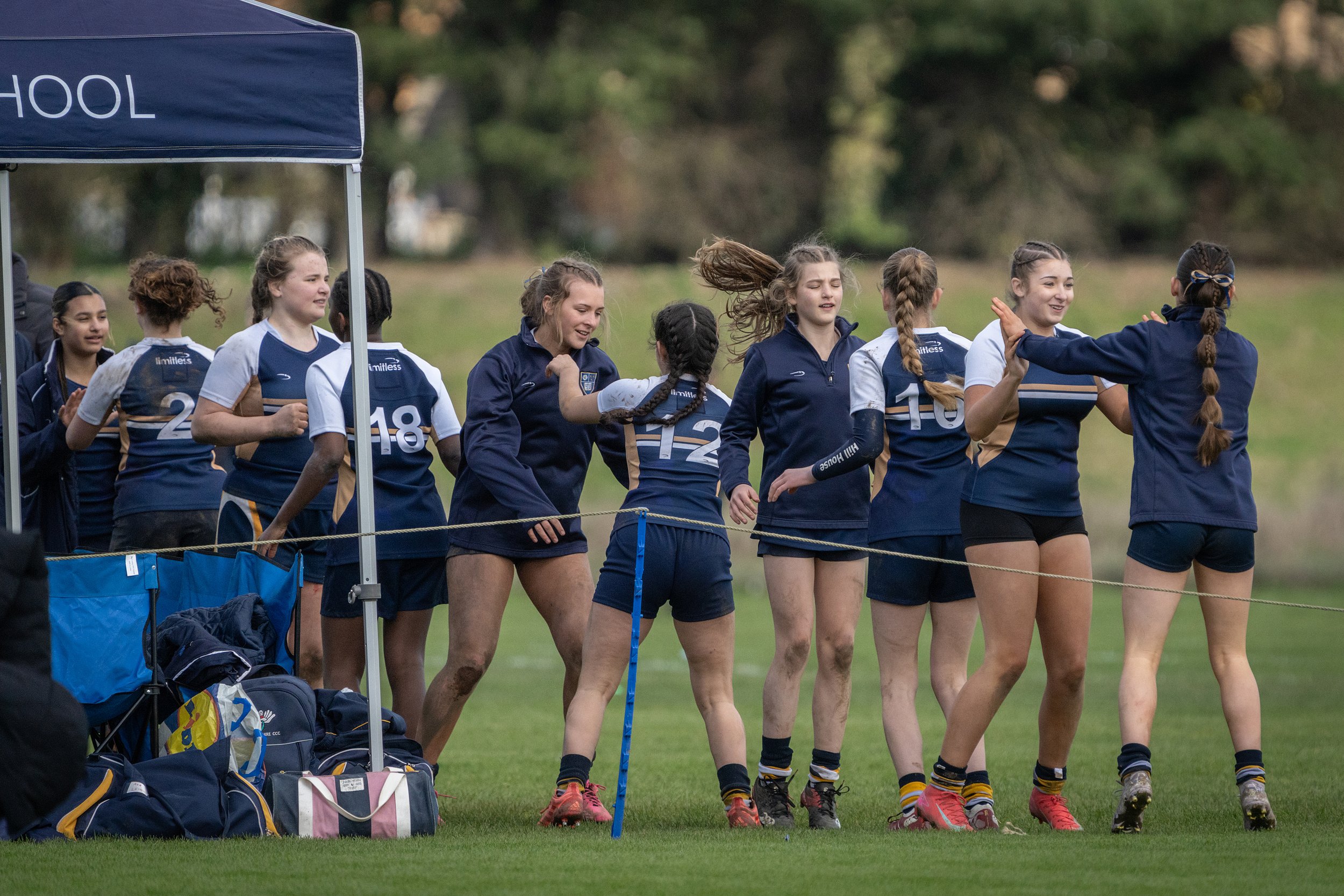 A group of female rugby players in navy and white uniforms standing together on a field during a game, some high-fiving and laughing.