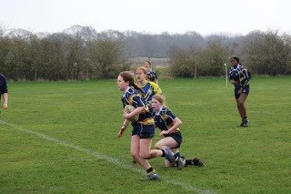 Girls playing rugby on a grassy field with trees in the background.