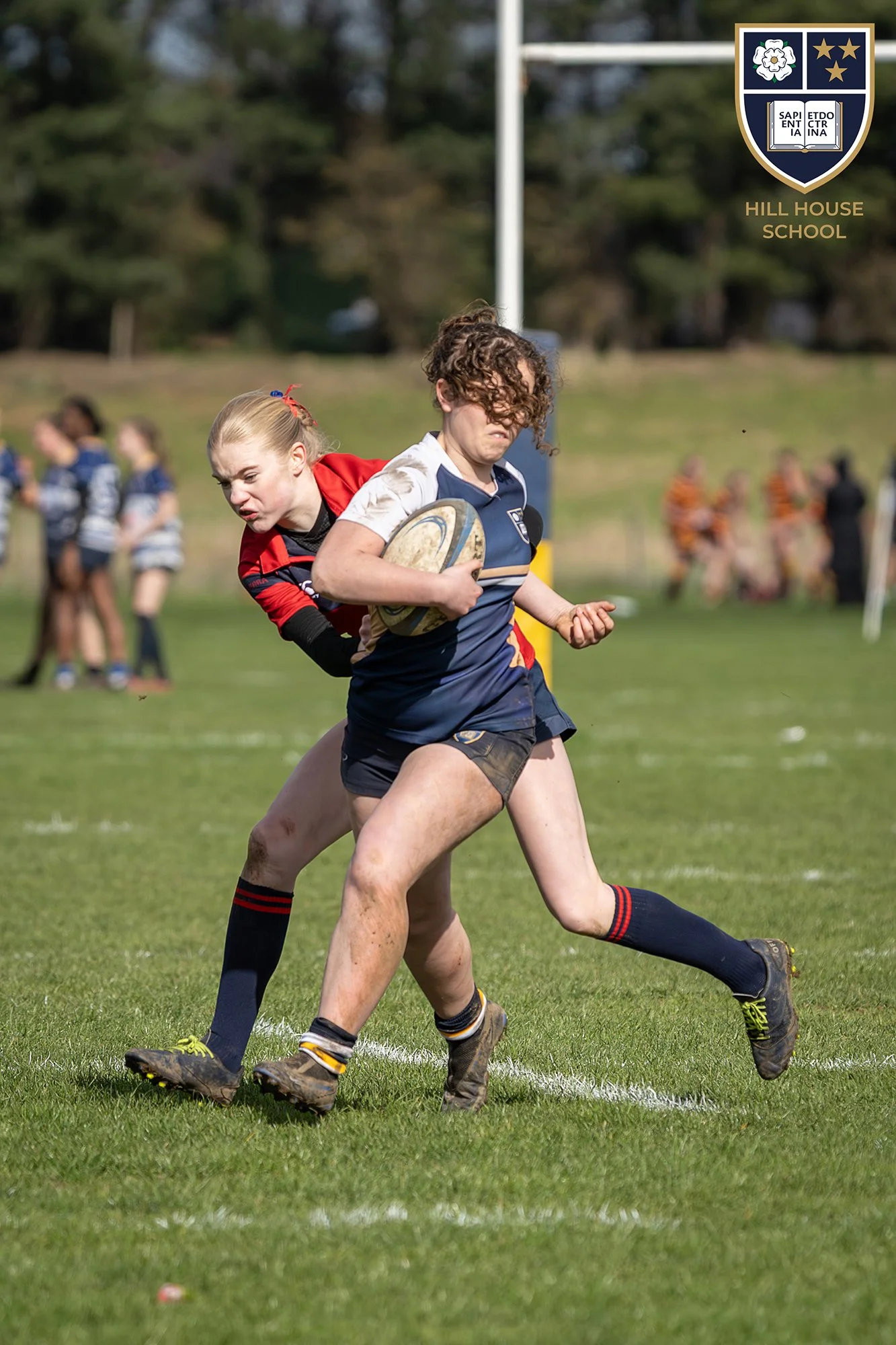 Two female rugby players in action, one carrying the ball while the other attempts to tackle her on a grassy field, with a school crest and the name "Hill House School" in the upper right corner.