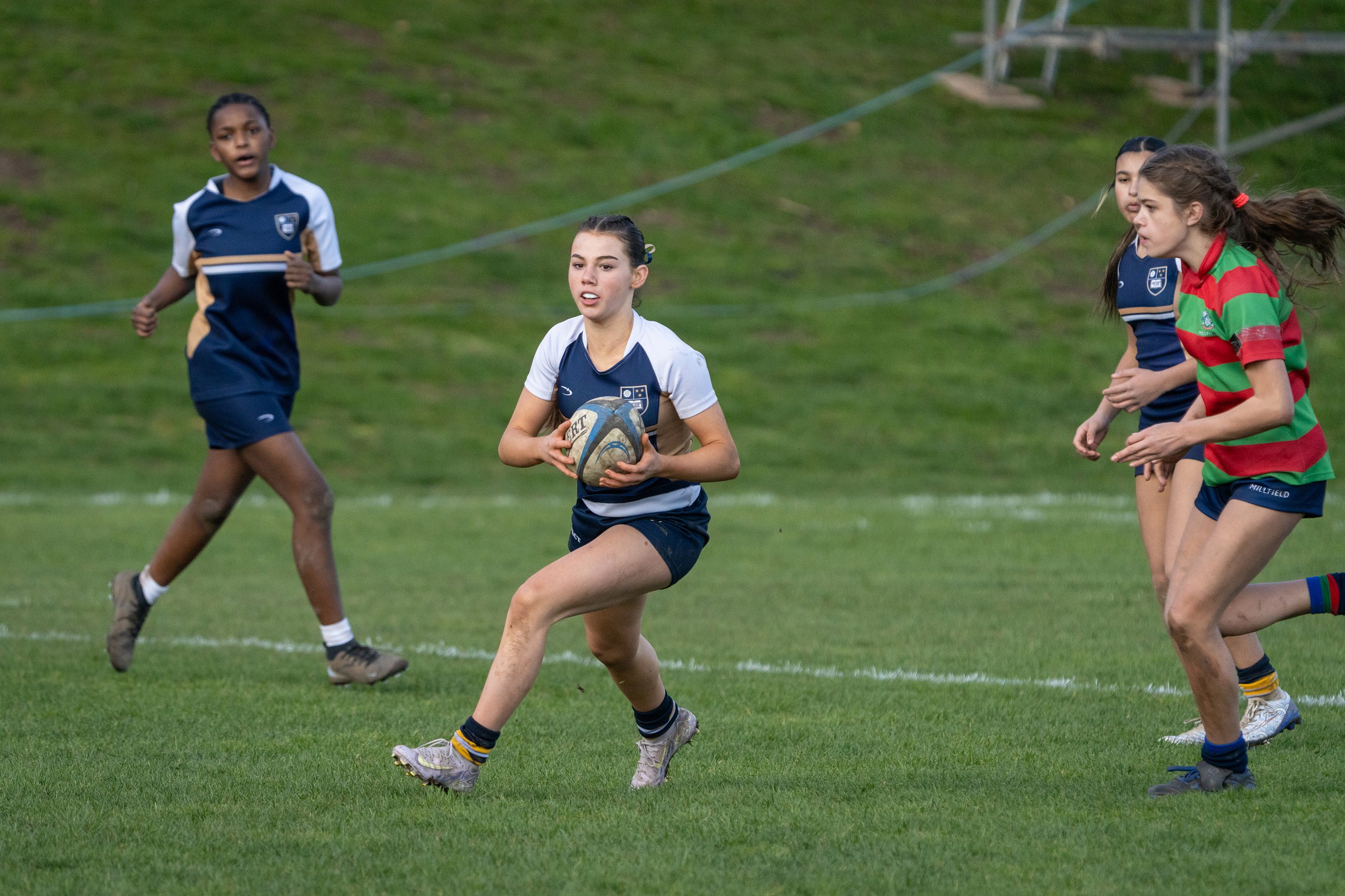 Young female rugby players in blue and red striped uniforms run on a grassy field during a game, with one girl holding a rugby ball.