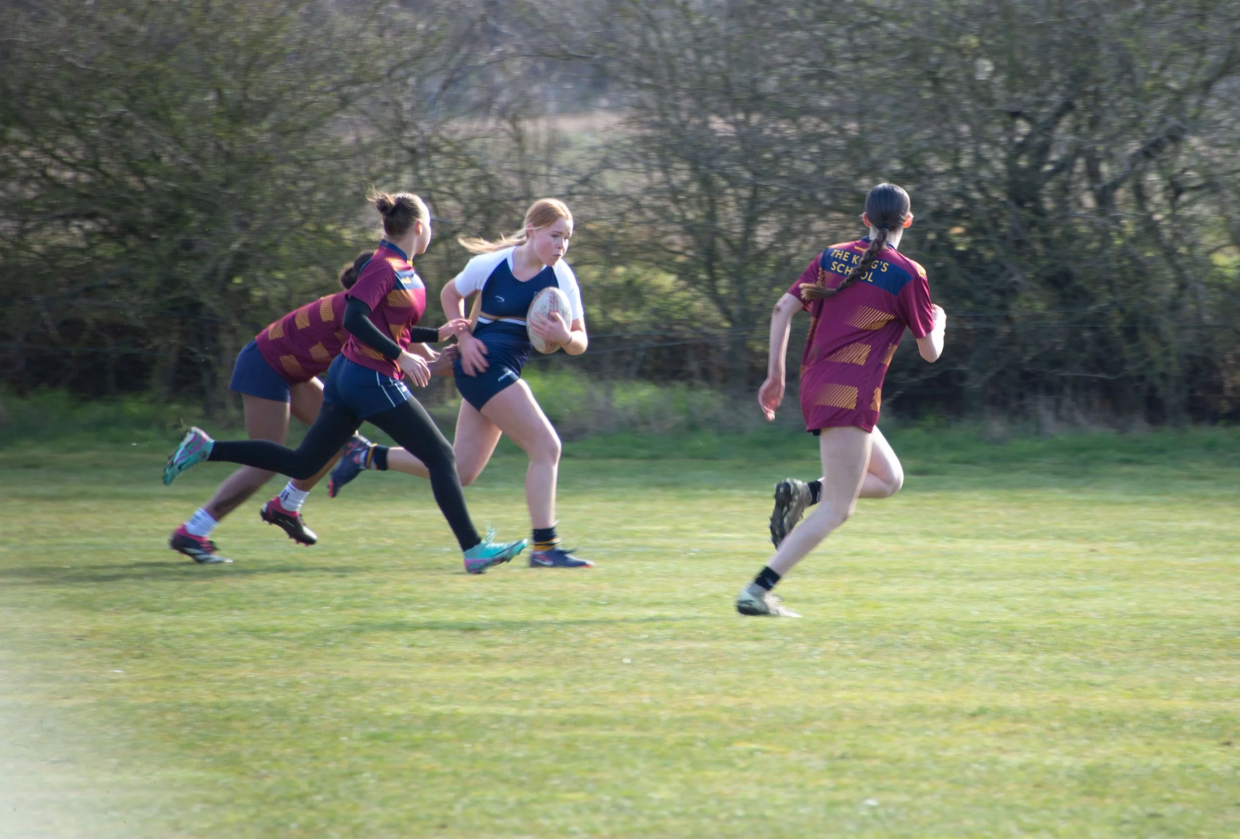 Young girl holding a rugby ball running on a field while two other girls chase her during a rugby game.