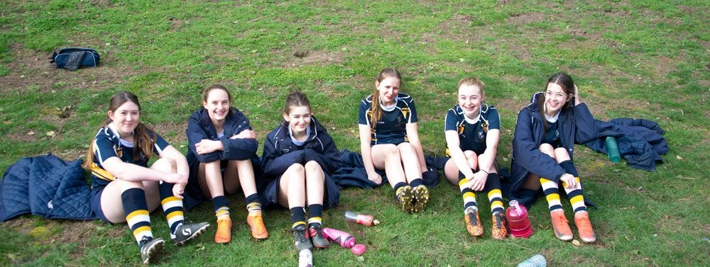 Six girls in sports uniforms sitting on grass with water bottles and jackets nearby.