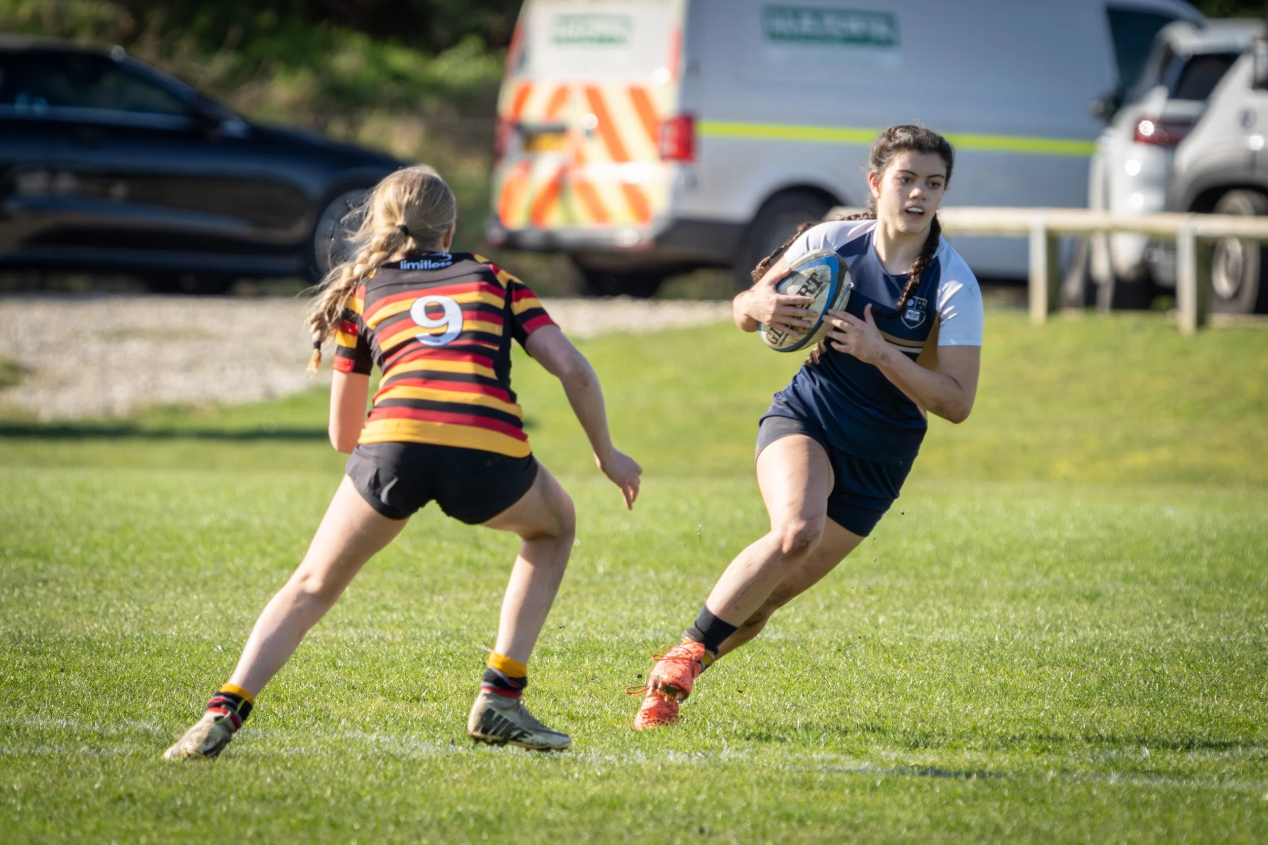 Two young women playing rugby on a grassy field, one running with the ball and the other chasing, with parked cars and a police vehicle in the background.