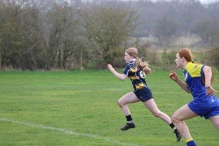 Two female rugby players running on a grassy field. One has blonde hair and is wearing a black and yellow uniform, the other has red hair and is wearing a blue and yellow uniform.