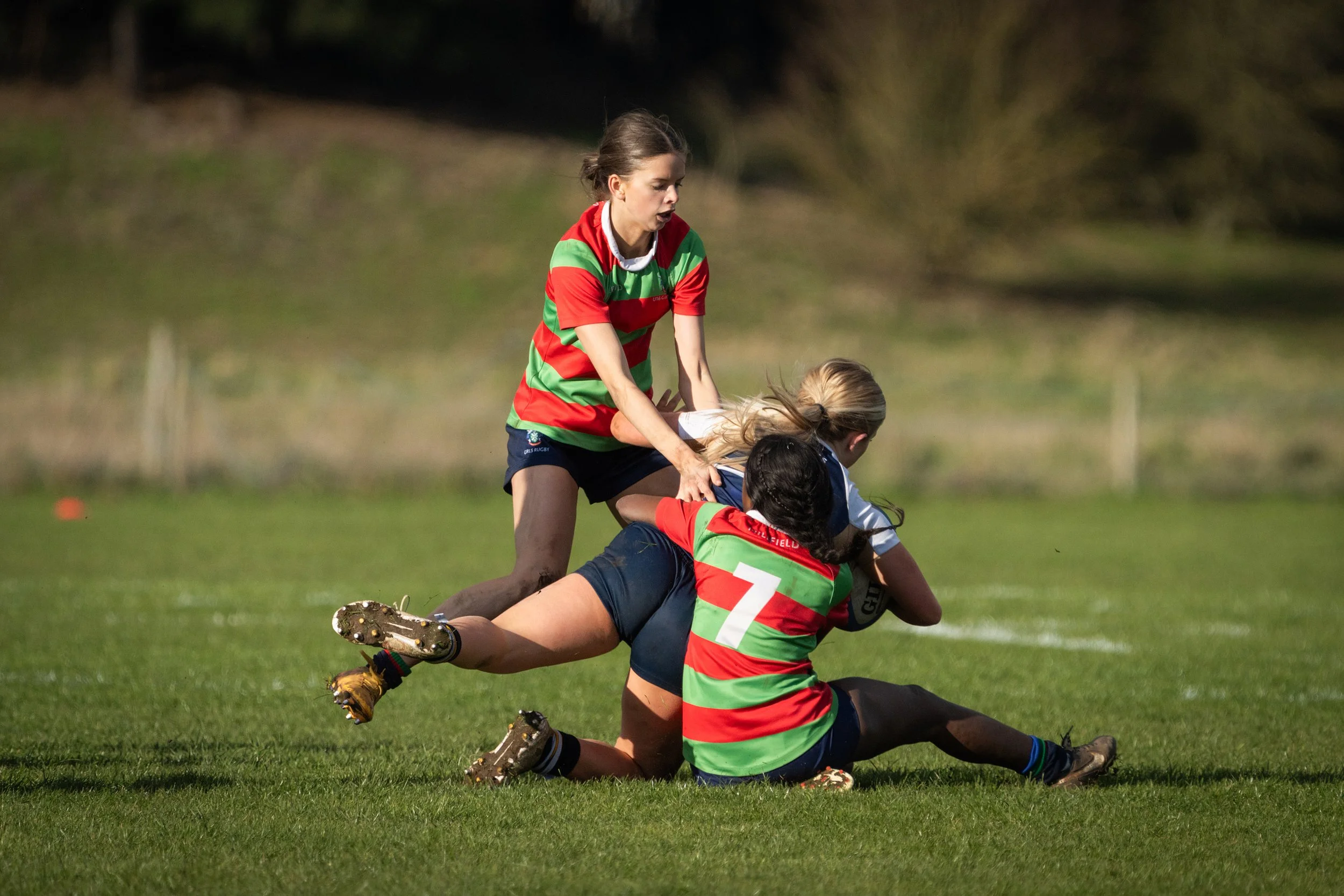 Girls playing rugby on a grassy field, one girl in a red and green striped uniform holding another girl in a blue and white uniform, both on the ground in a tackle, with a background of blurred trees.