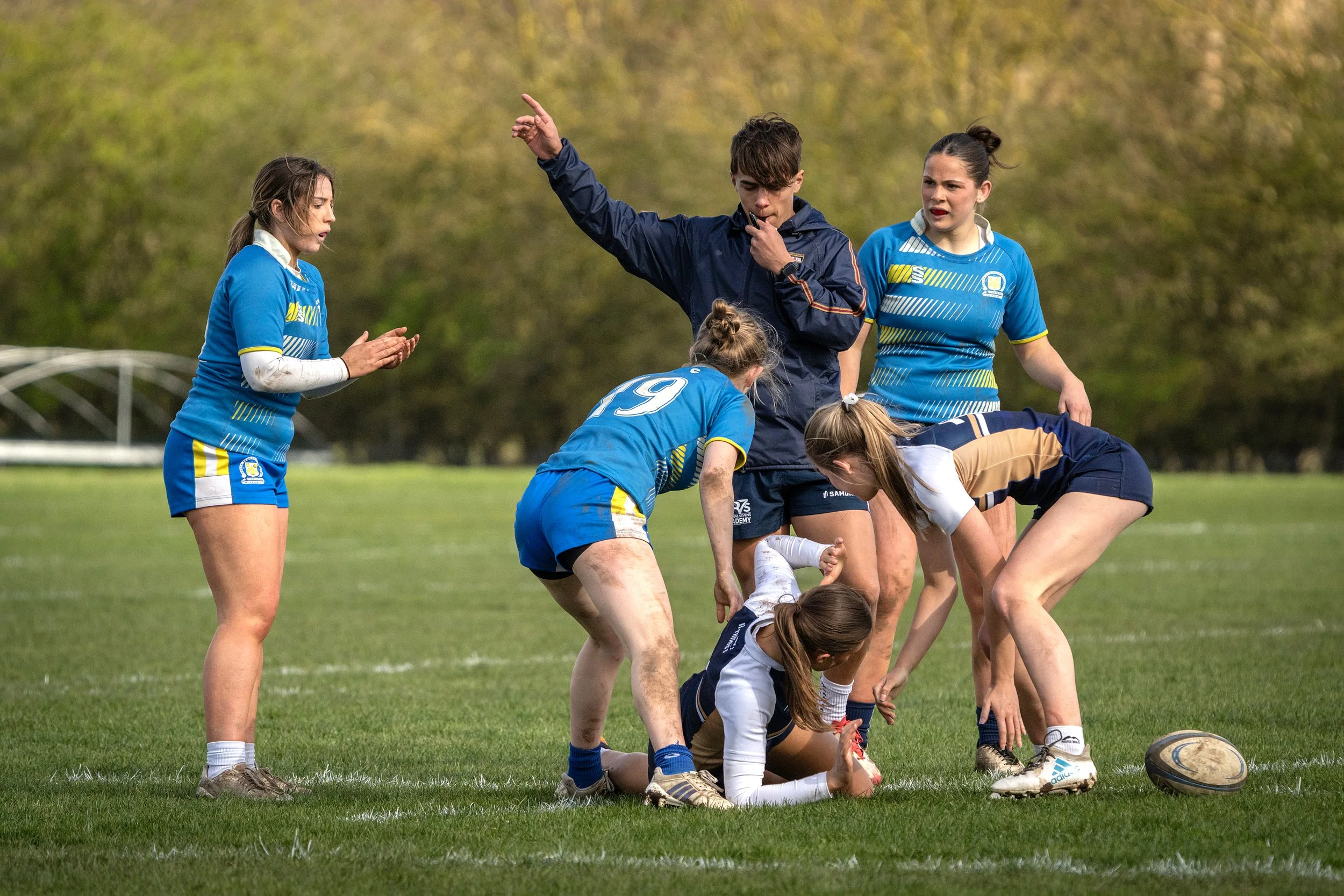 Women rugby players in blue and white uniforms are on a grassy field, participating in a game or practice. One player is lying on the ground while others are bending over her. A referee in a navy jacket is standing nearby, gesturing with his right ar