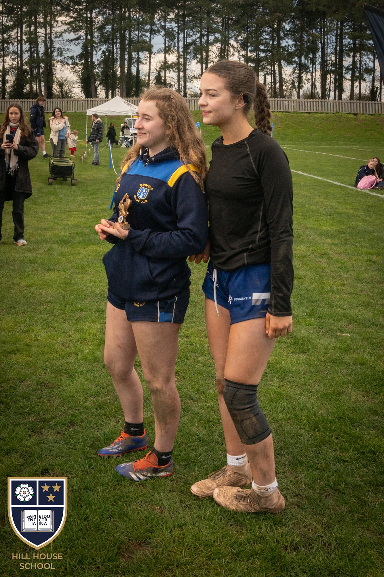 Two young women standing side by side on a grassy field, one in a blue sports jacket holding a small trophy, and both wearing athletic gear, with spectators and trees in the background.