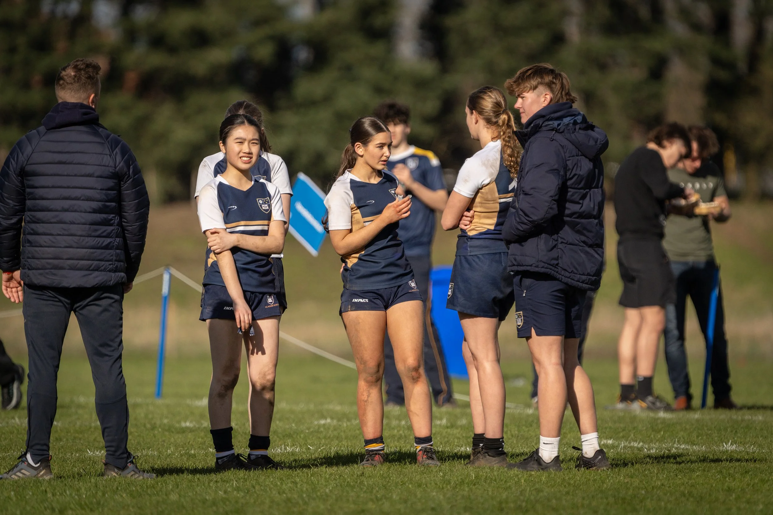 Group of young female and male rugby players in team uniforms standing on a grassy field having a conversation, with trees in the background.