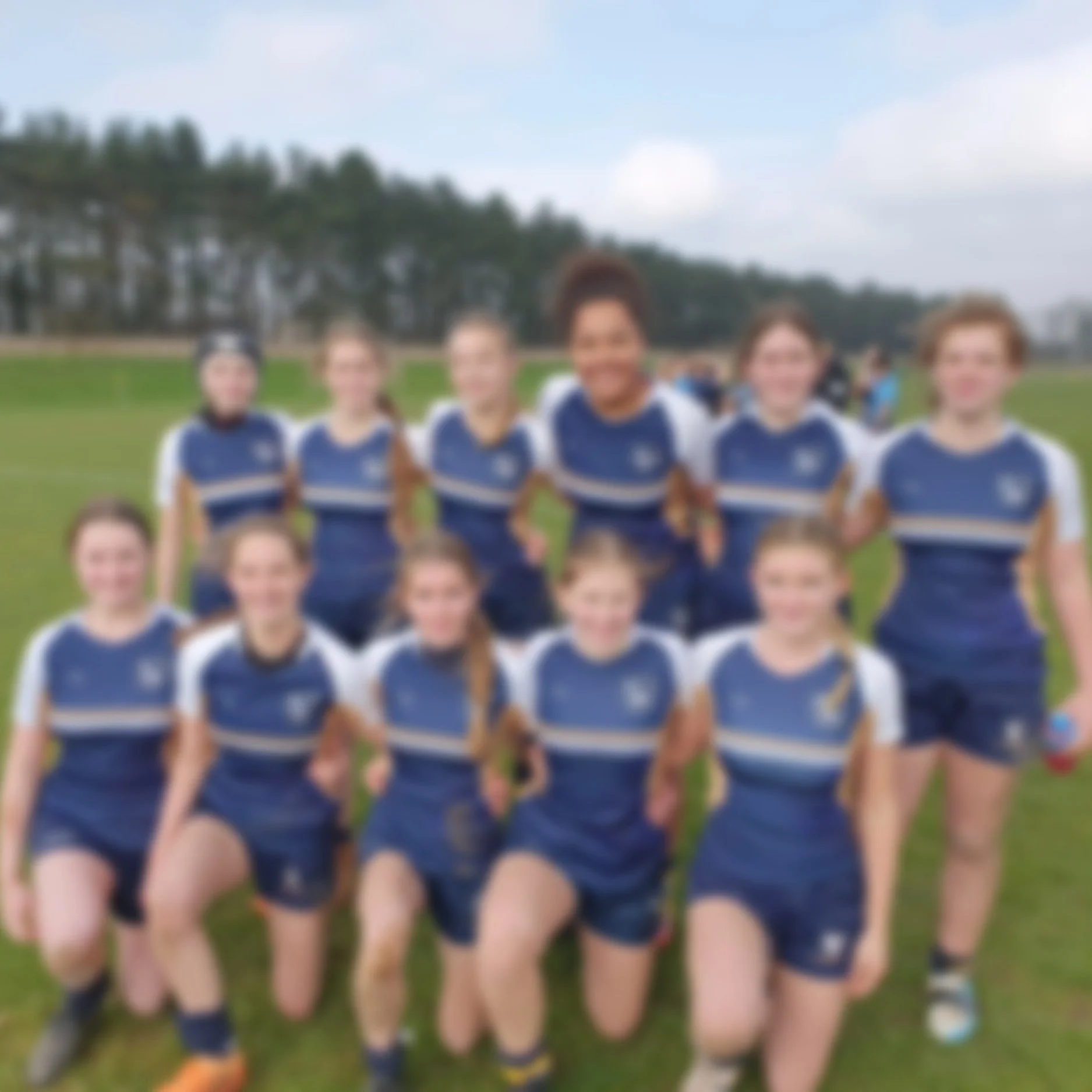 A photo of a group of young girls in blue sports uniforms standing outdoors on a grassy field, likely a soccer team, with trees and a cloudy sky in the background.