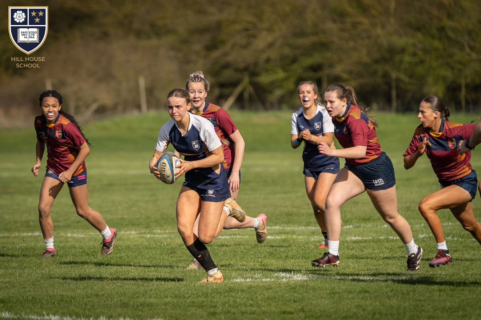 Girls playing rugby on a grassy field, wearing sports uniforms, with one girl holding a rugby ball and running forward.