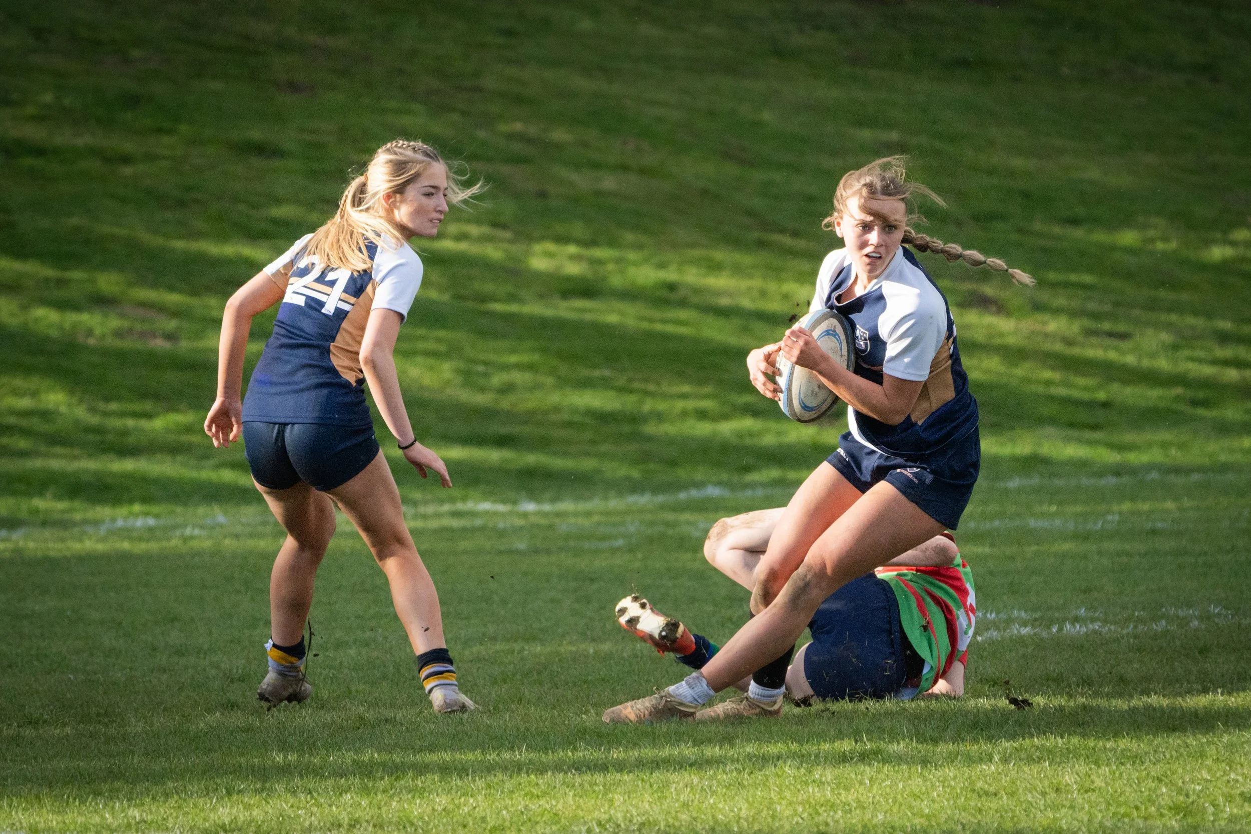 Two women playing rugby on a grassy field, one running with the ball, and the other trying to tackle her, both wearing athletic uniform.